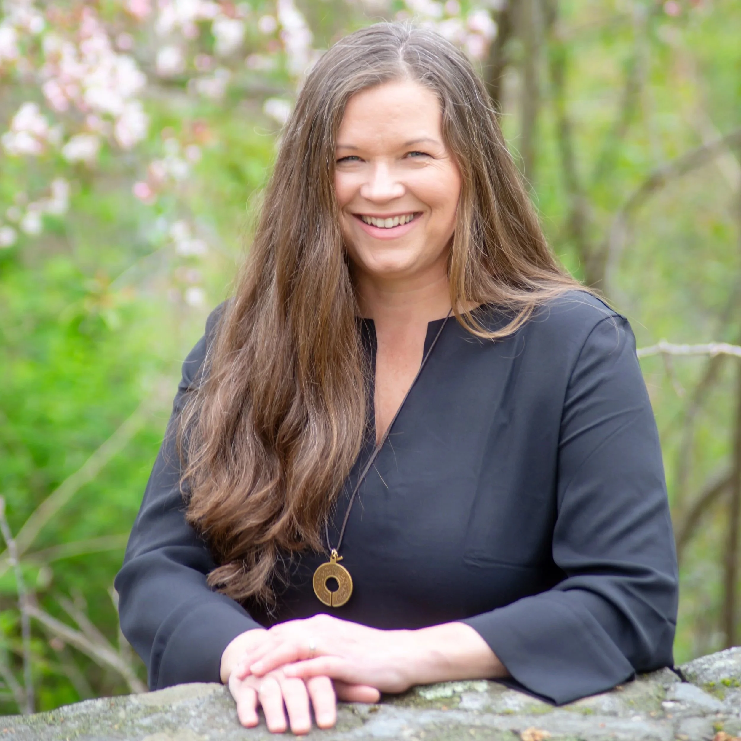 A woman with long brown hair, smiling, wearing a black jacket with a gold pendant, resting her arms on a stone surface outdoors with blurred greenery in the background.