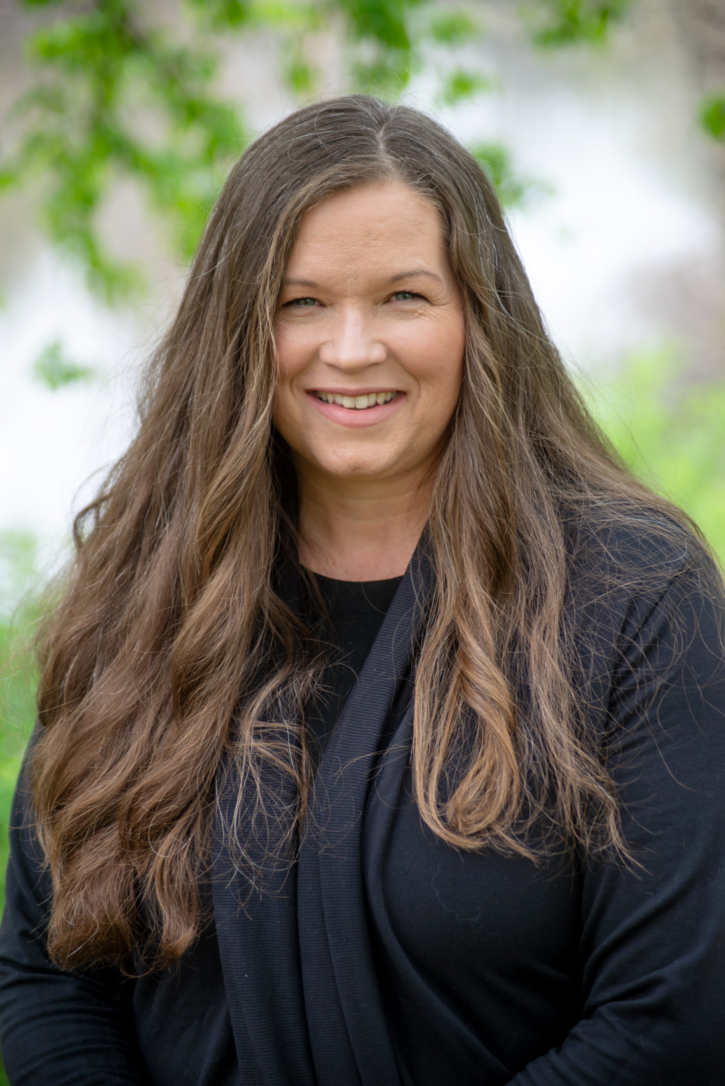 Close-up portrait of a woman with long wavy brown hair, smiling, outdoors with blurred green foliage background.
