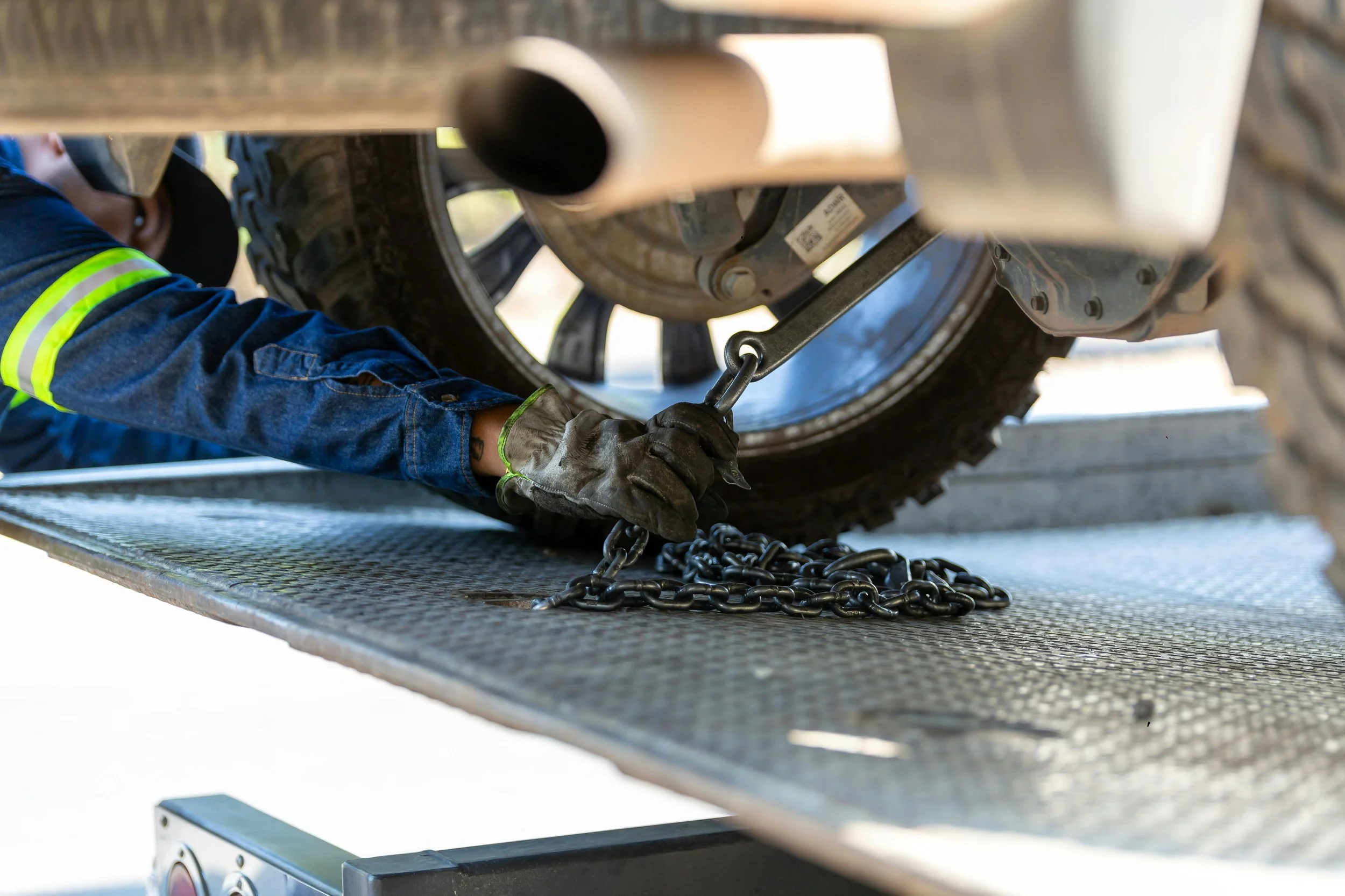 A worker wearing gloves and a denim shirt with yellow reflective stripes is securing a car tire with a chain while lying on a metal platform.