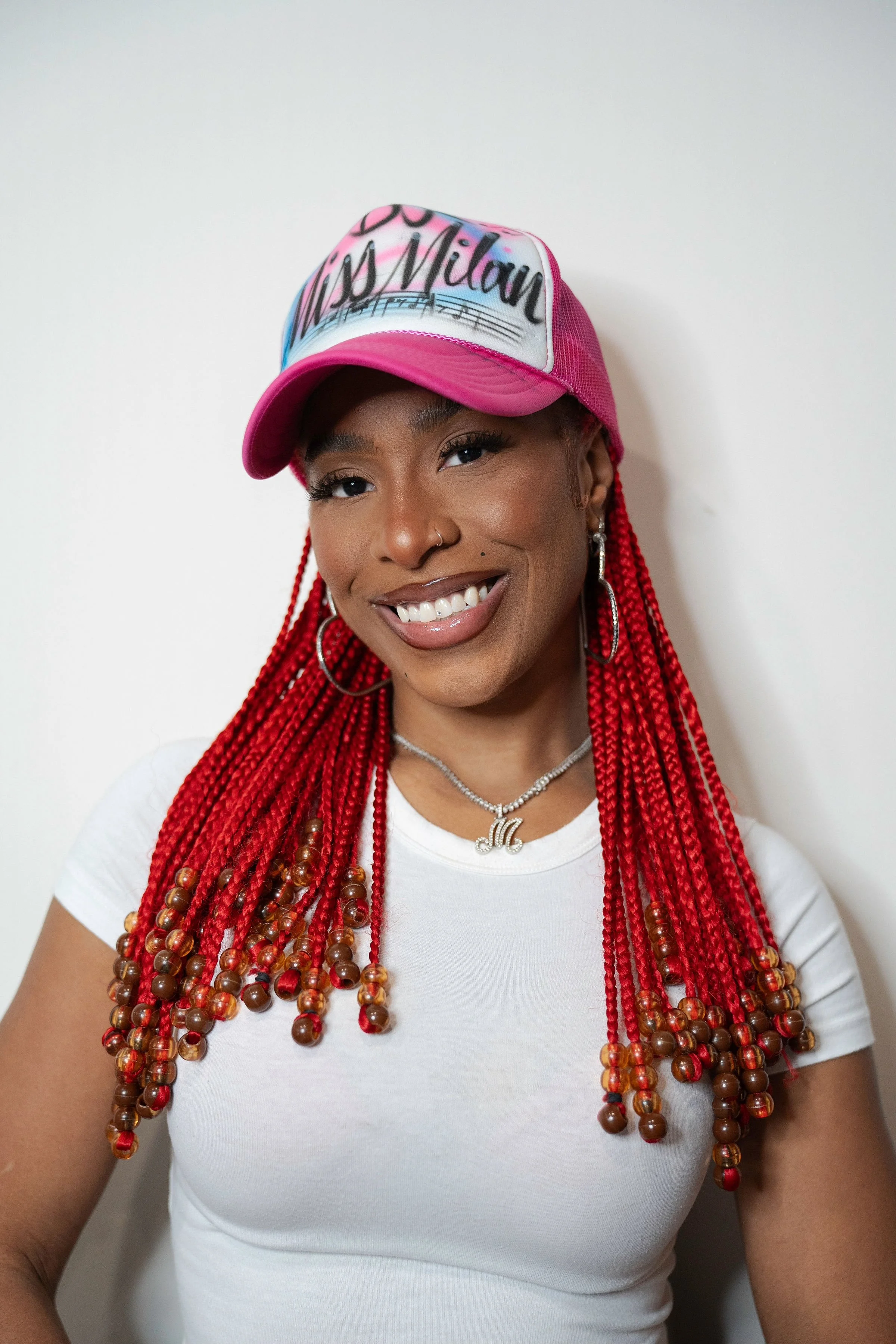 Headshot image of Dj Miss Milan smiling. She has red braids with beads and a pink airbrushed hat.