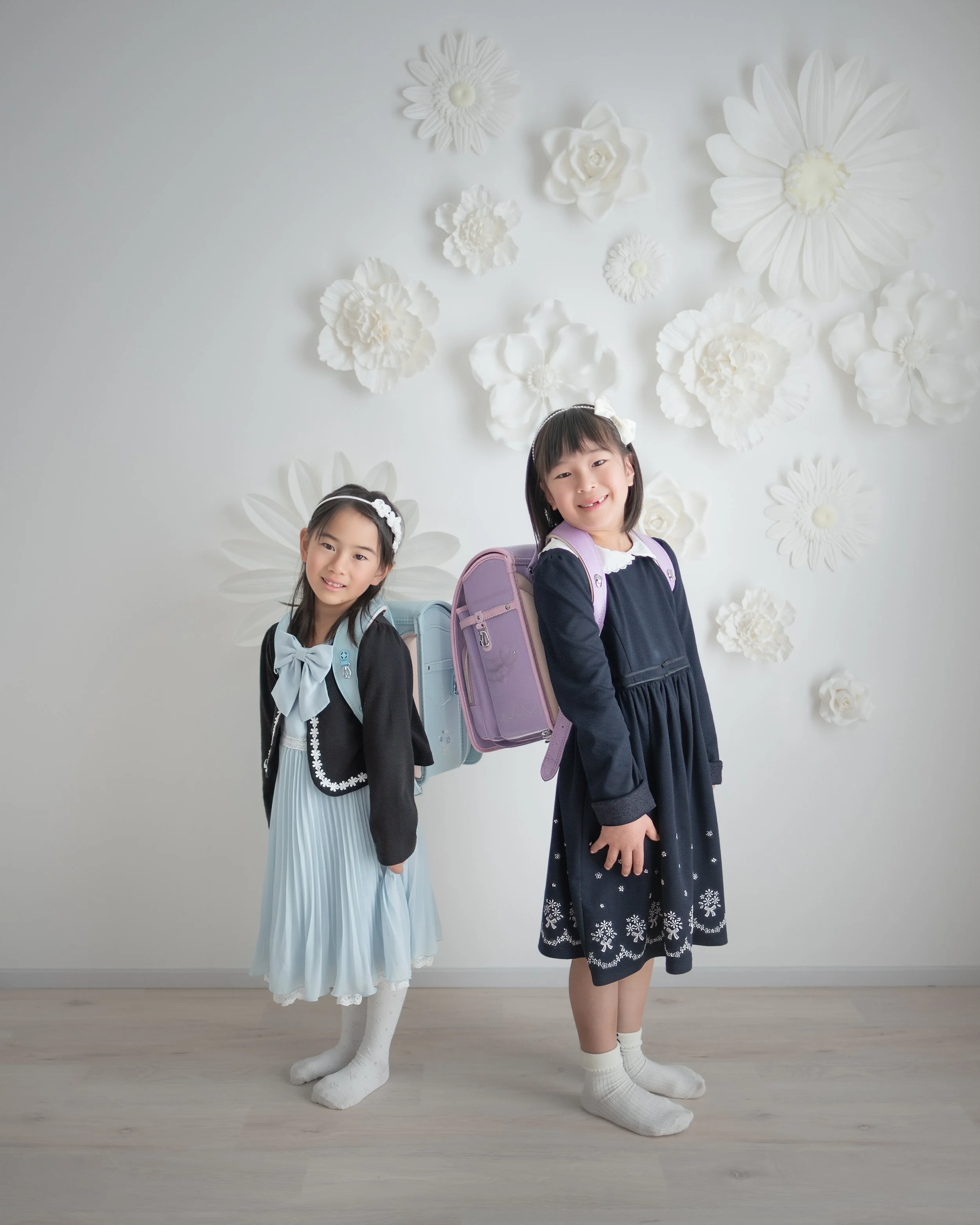 白い背景に白い花の装飾が配置された壁の前で、正装した少女二人が笑顔で立っている写真