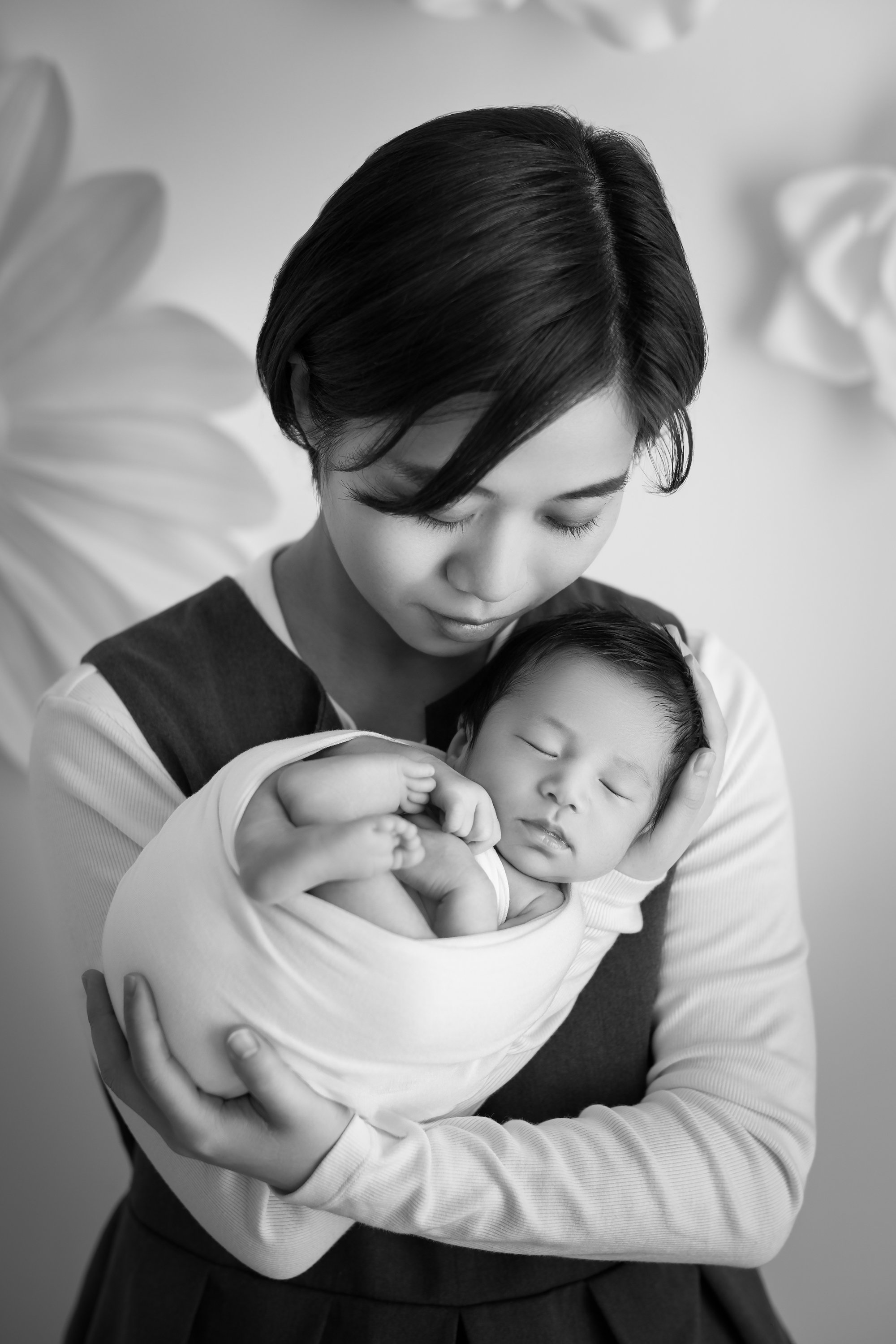 母親が赤ちゃんを抱いている白黒の写真。赤ちゃんは眠っている。背景に花の飾り。