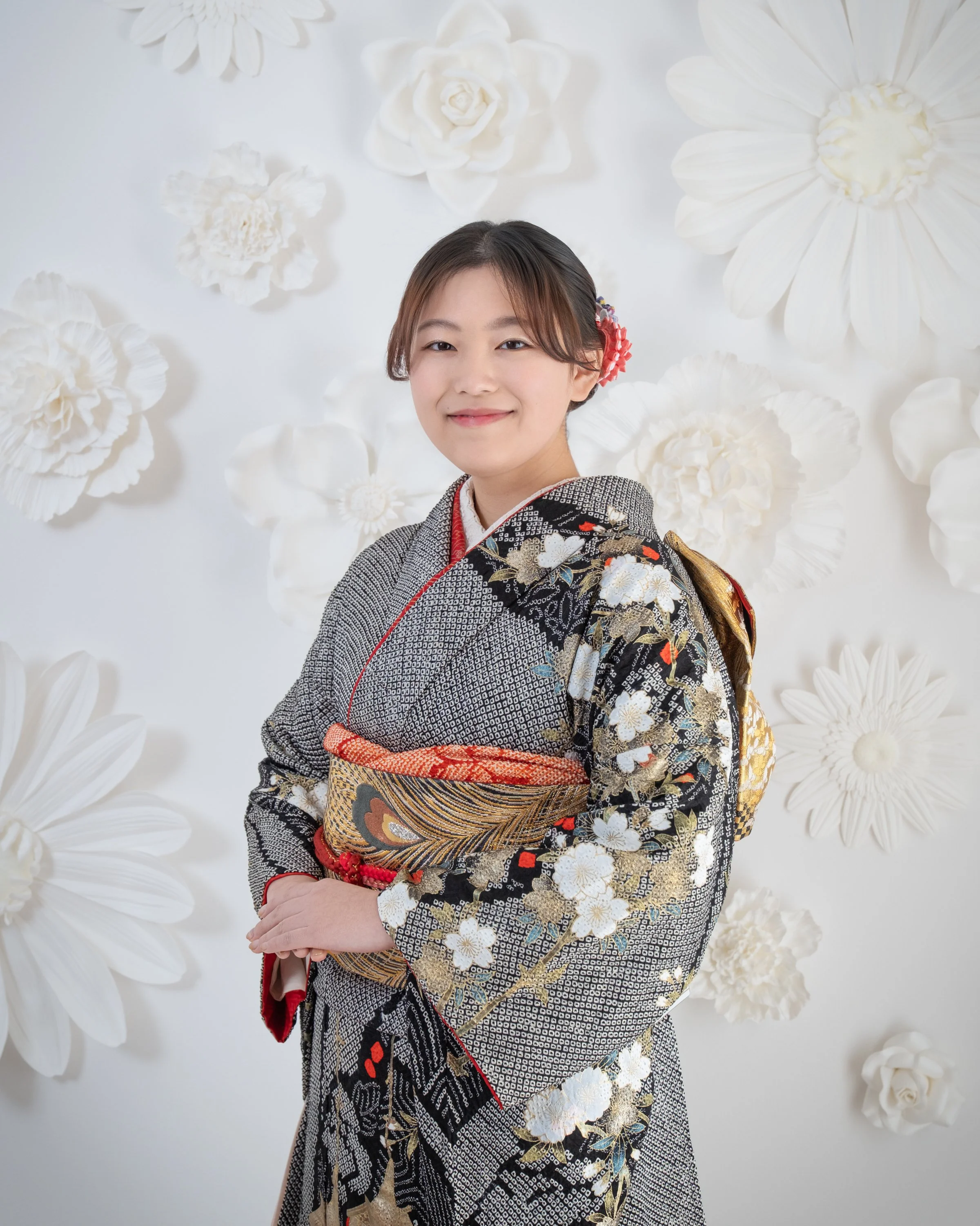 白い壁に黒、白と金の着物を着た若い女性が微笑んでいる写真。