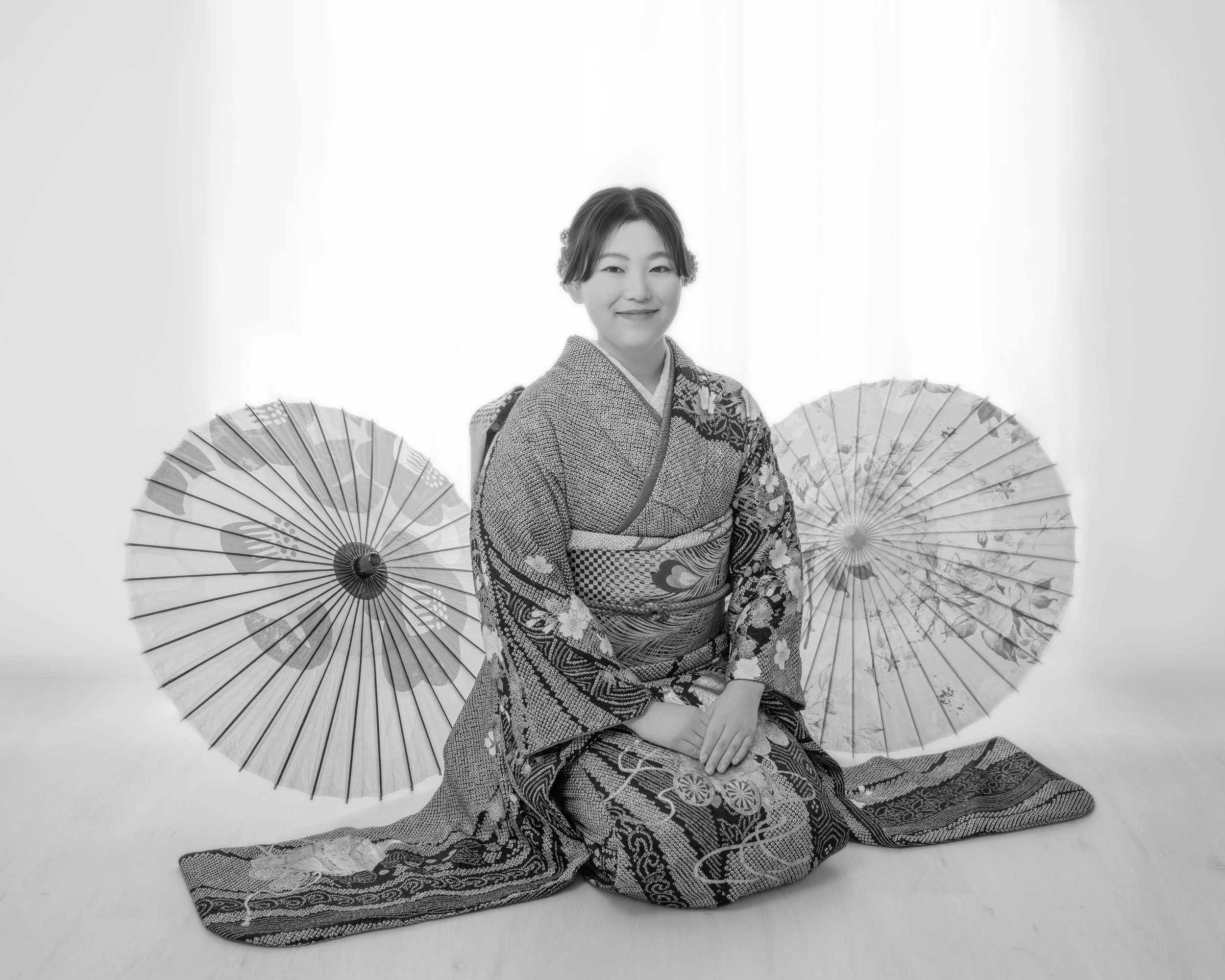 着物を着た女性のポートレート。二つの和柄の傘の間に座り、にこやかに微笑んでいる。背景は白い。