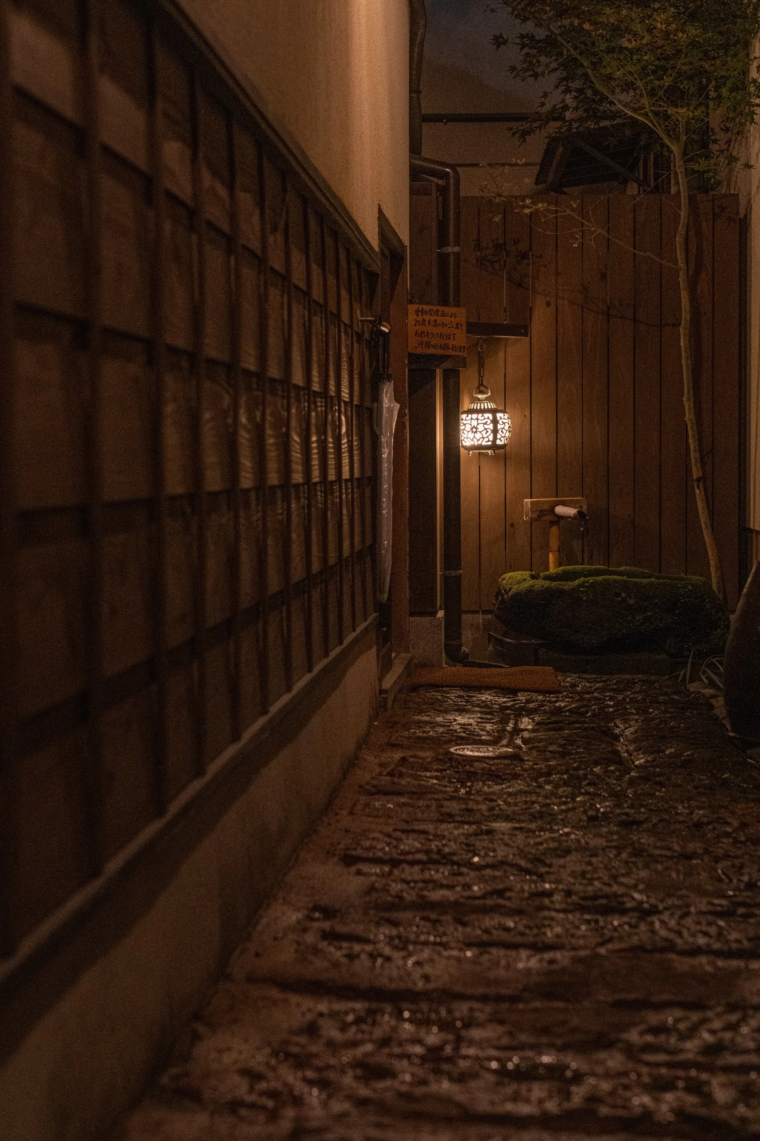 暗い夜の居酒屋の通路。木製の壁と石畳の道、ランタンの灯りが見える。