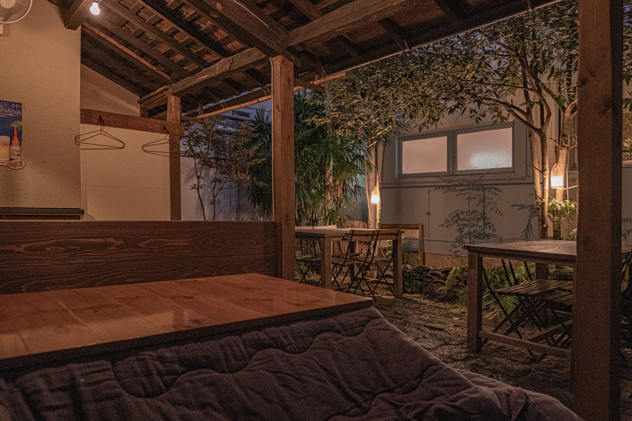 木製のテーブルとチェアが配置された屋外の居心地の良いテラス、夜景が見える窓と植物、暖かな照明。