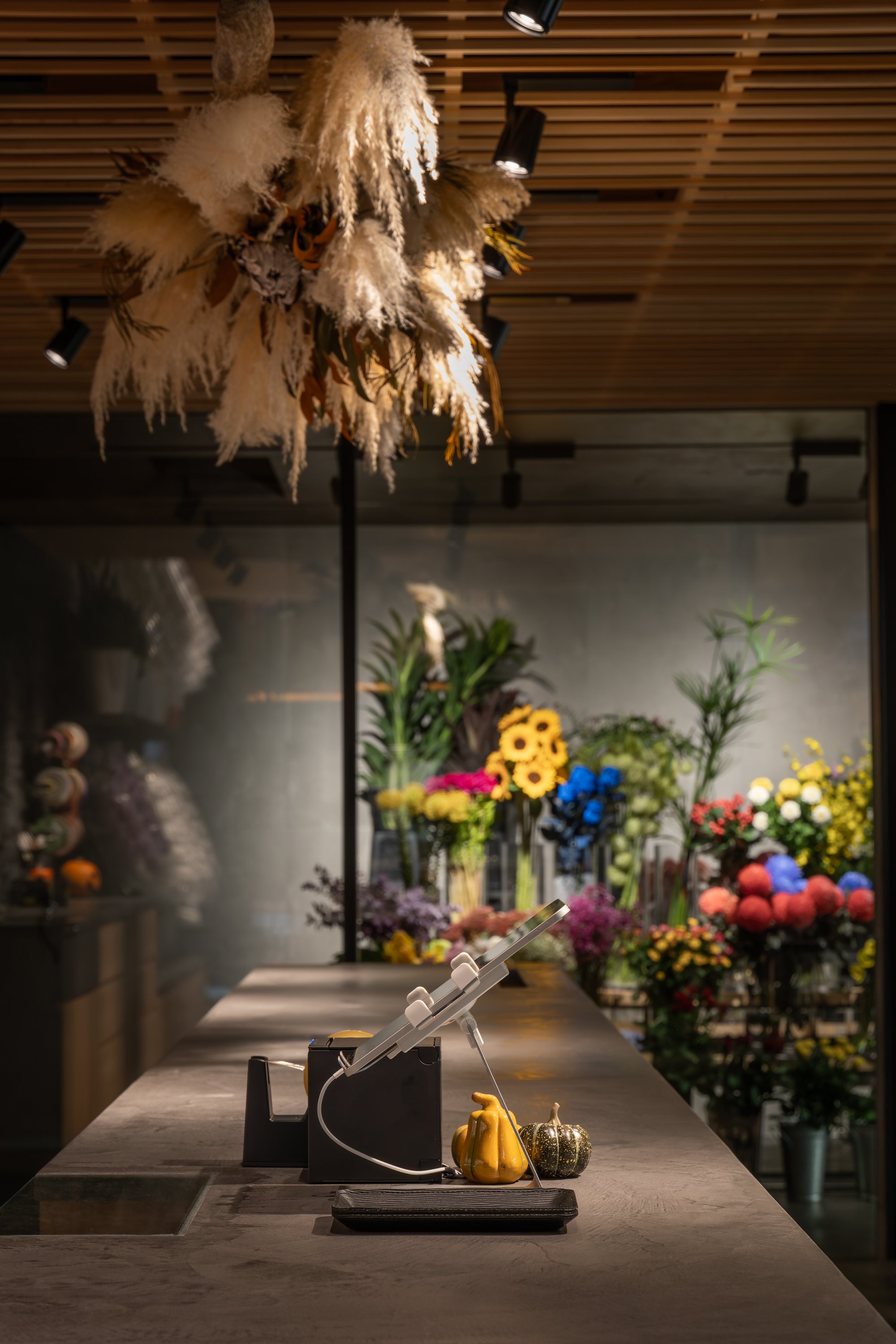 花屋のカウンターに黄色と緑の小さなかぼちゃの置物とレジのタブレットが置かれている。背景には色とりどりの花束が飾られている。天井には大きなドライフラワーのアレンジメントが吊るされている。