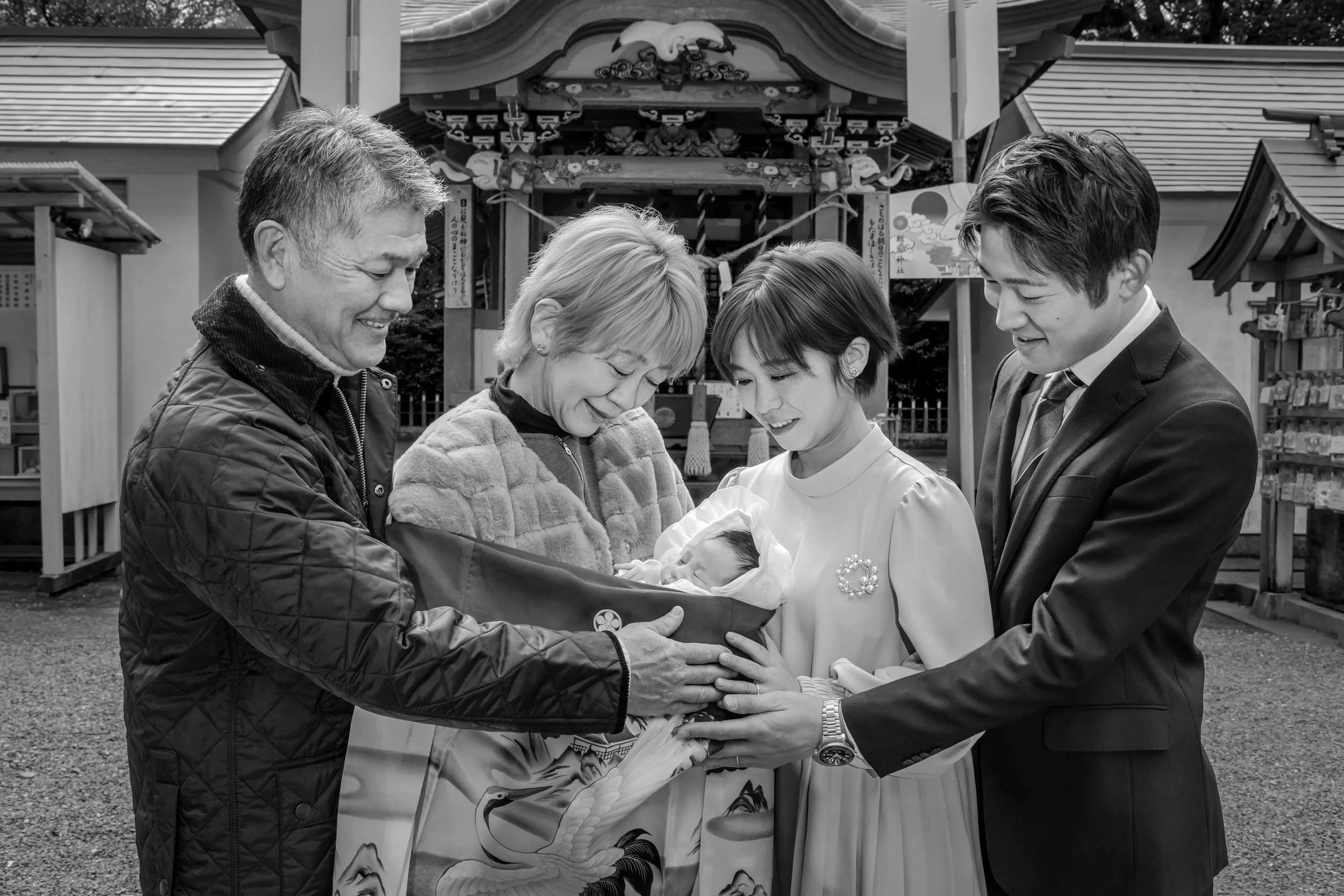 お宮参りの家族の写真。赤ちゃんをみんなで見ている。背景には神社の社殿が見える。