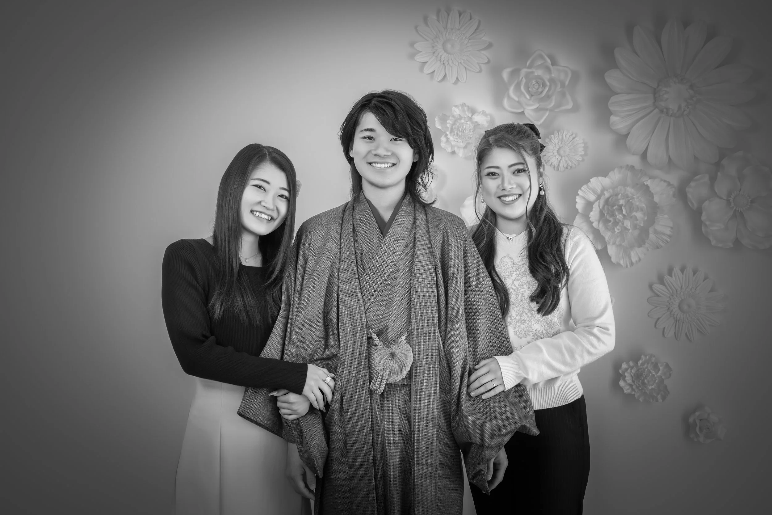 和装の弟と洋装の姉二人が笑顔で並んでいる写真。背景には花の装飾がある。