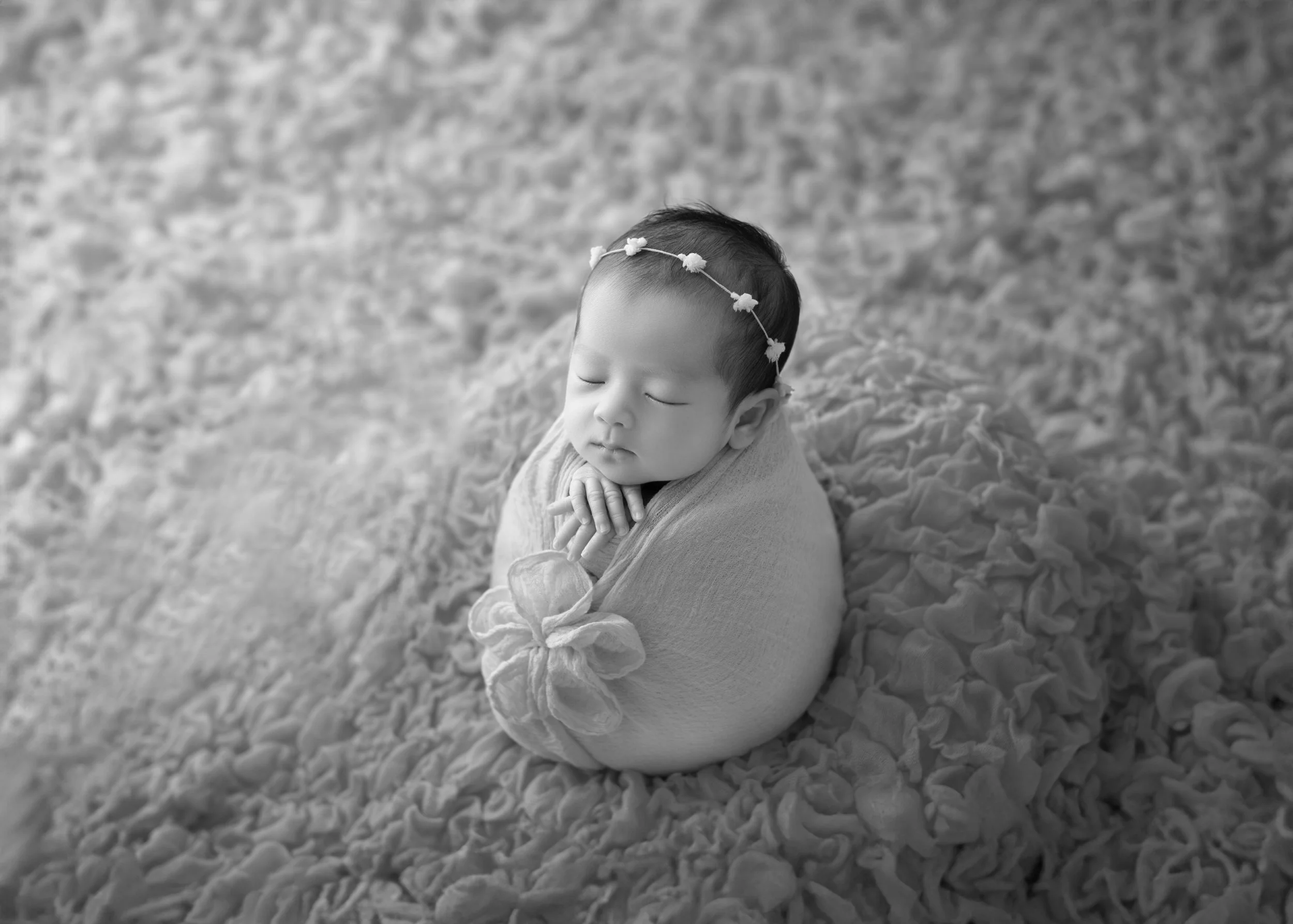 赤ちゃんが眠っている写真。髪飾りを付けて、スワドリングされた状態で、柔らかい布で包まれている。ベッドのようなふわふわの寝具の上に横たわっている。