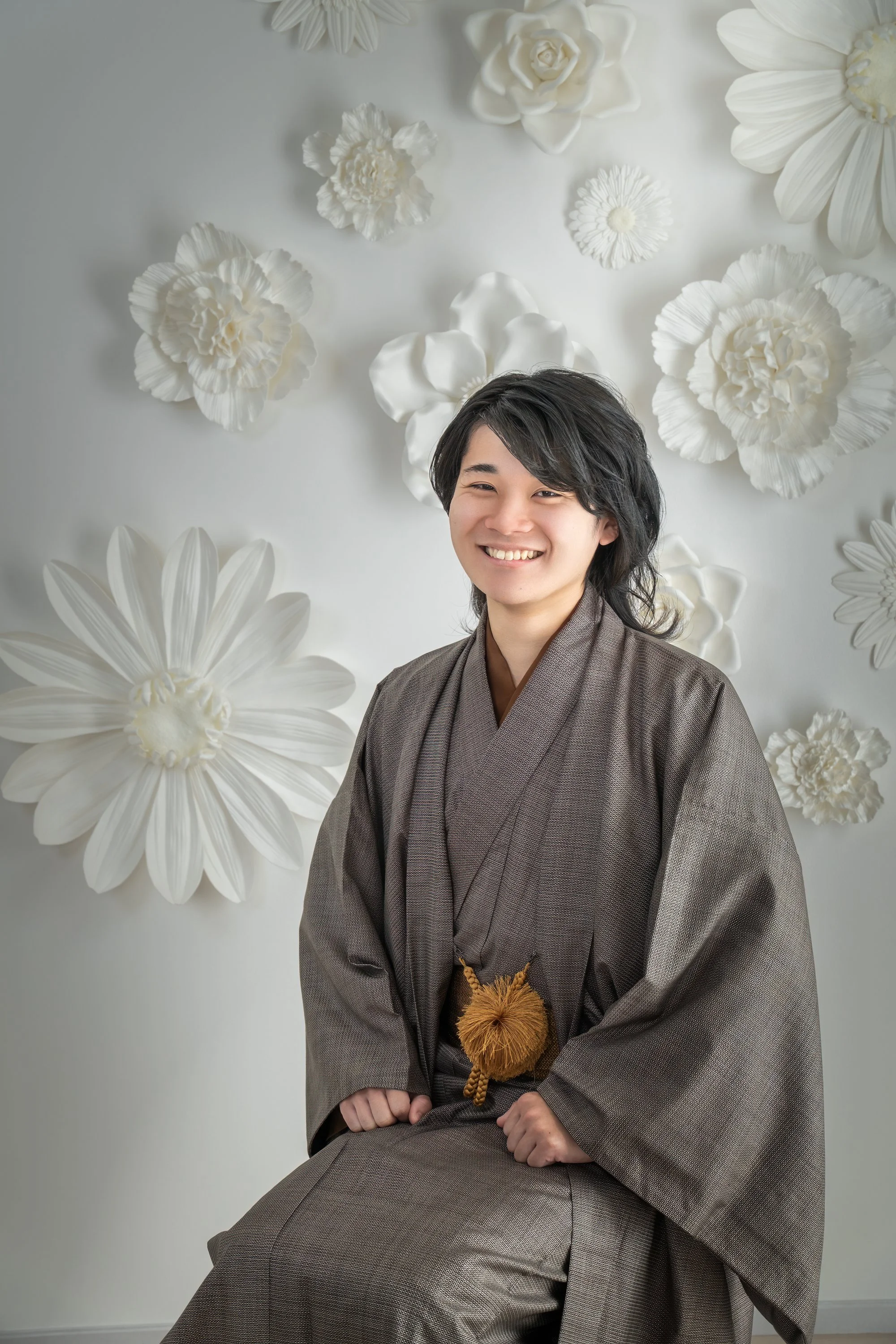 着物を着た男性のポートレート。背景には大きな白い花の装飾。男性は笑顔で座っている。