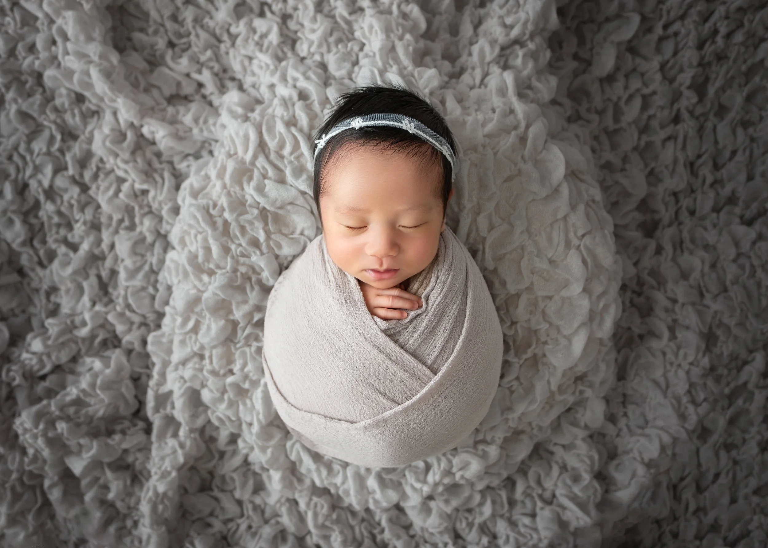 クリーム色の柔らかい布団に包まれた眠る赤ちゃん。頭に白いリボンのついたヘアバンドをつけている。