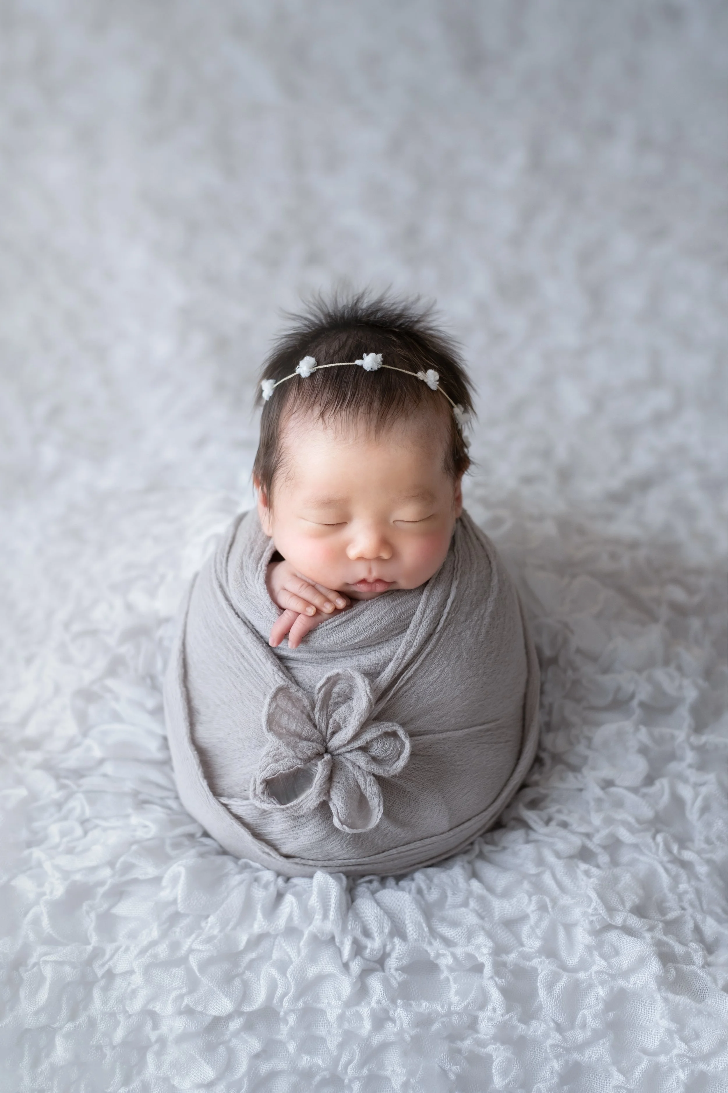 灰色の布に包まれた眠っている赤ちゃん、花の形の飾りがついたリボンをつけている