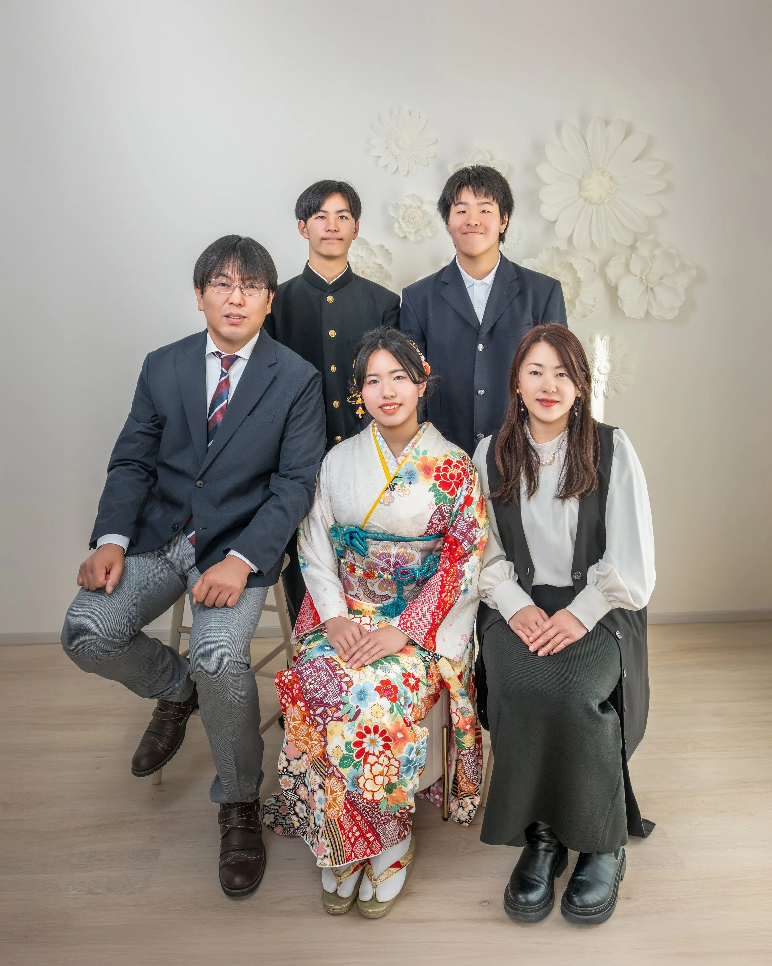 和服を着た女性とビジネススーツの男女が室内でポーズをとっている家族写真。背景に白い花の装飾がある。