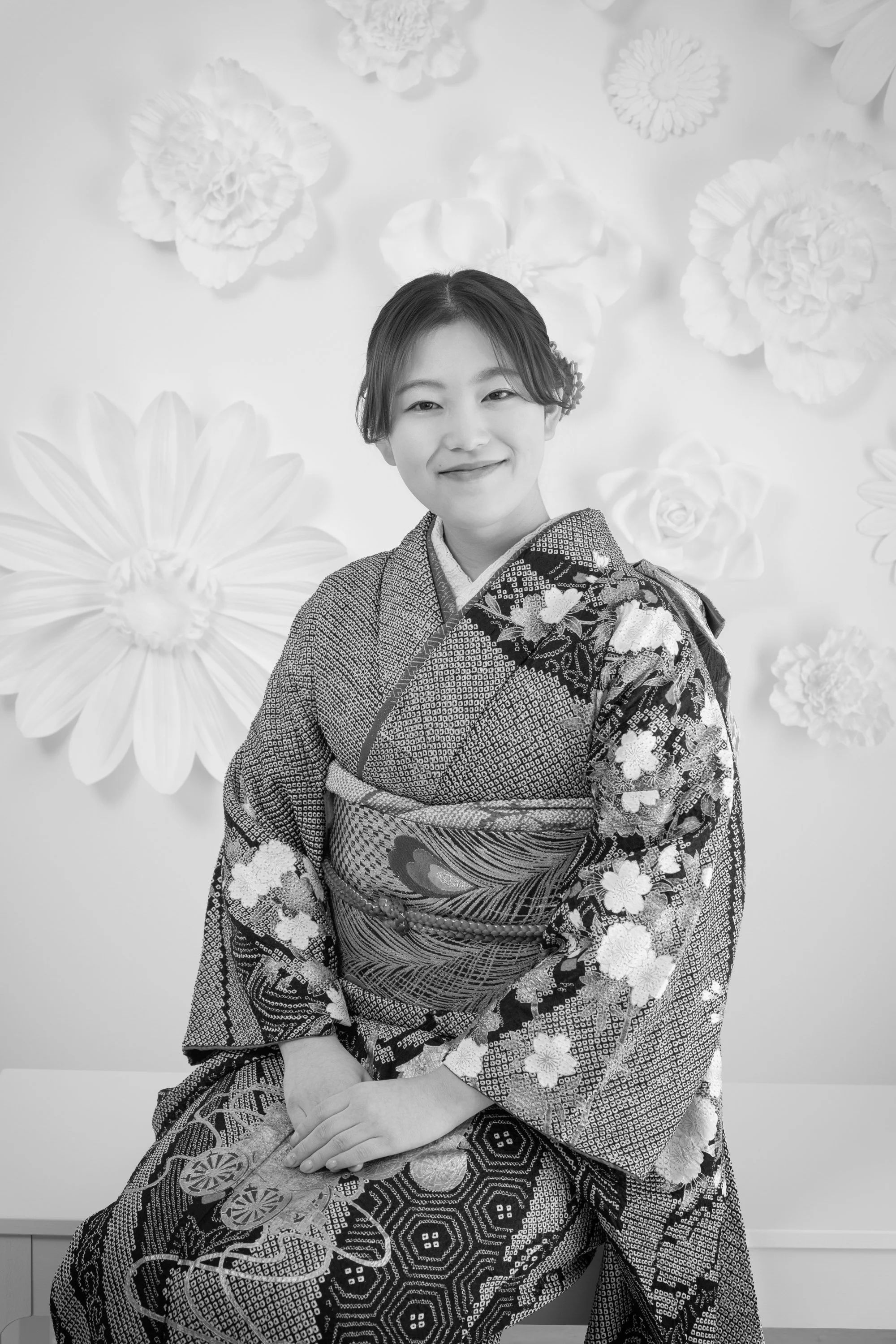 着物を着た若い女性が笑顔でポーズをとっている写真。背景には大きな花の装飾がある。