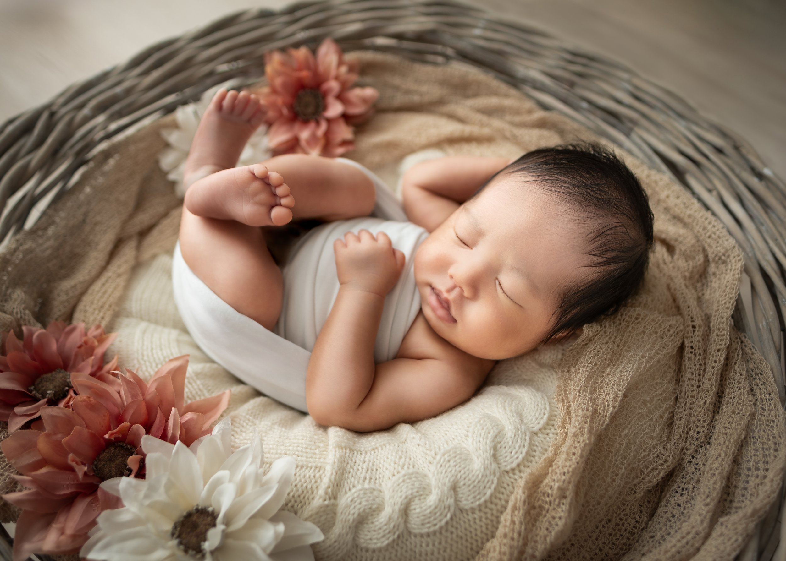 布で包まれた赤ちゃんが丸いバスケットの中で眠っている。周りに花が飾られている。