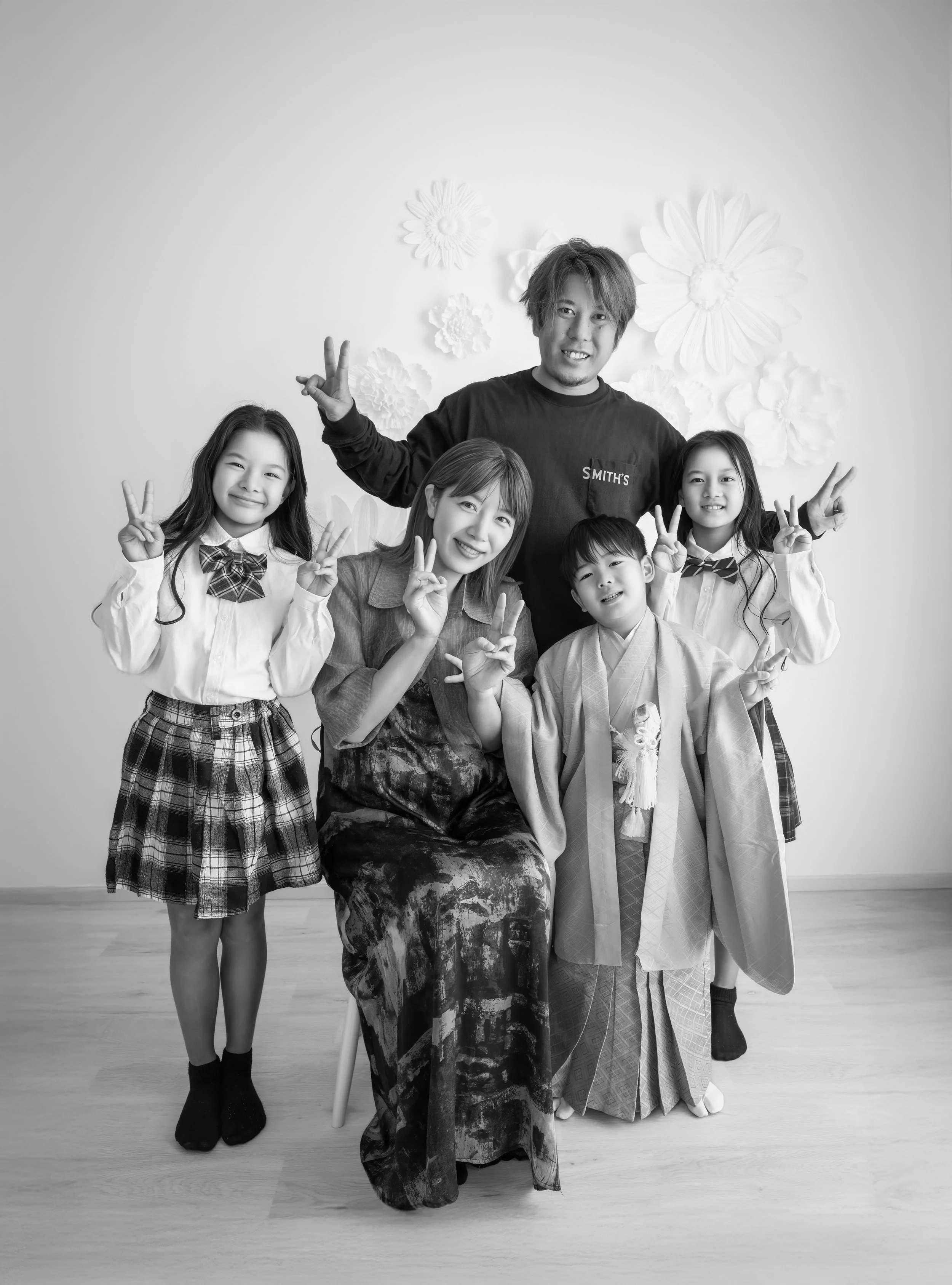 家族が和服と洋服でポーズをとって笑顔で写真を撮っています。背景には花の装飾があり、みんな手でVサインをしています。