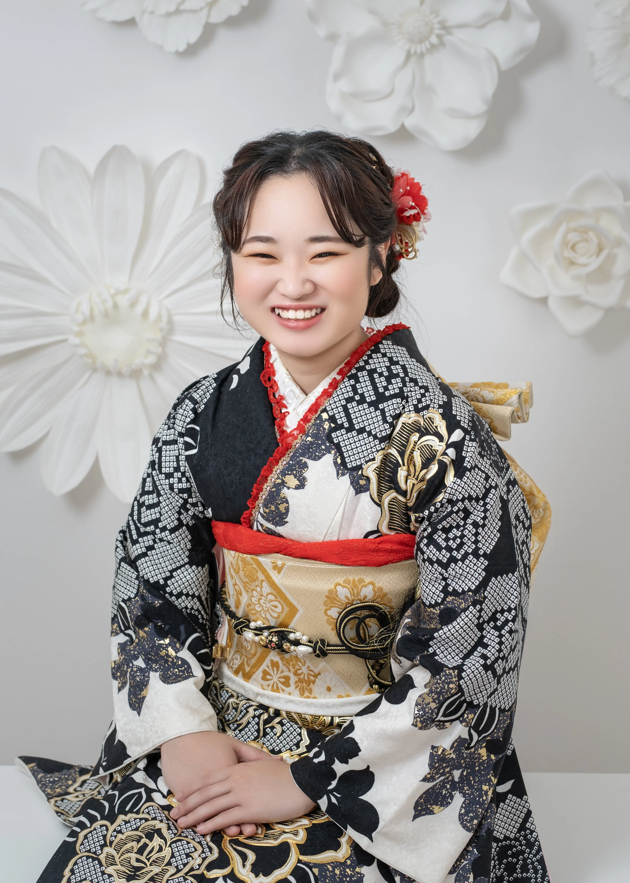 笑顔の女性が着物を着て座っている写真。背景に大きな白い花の装飾がある。