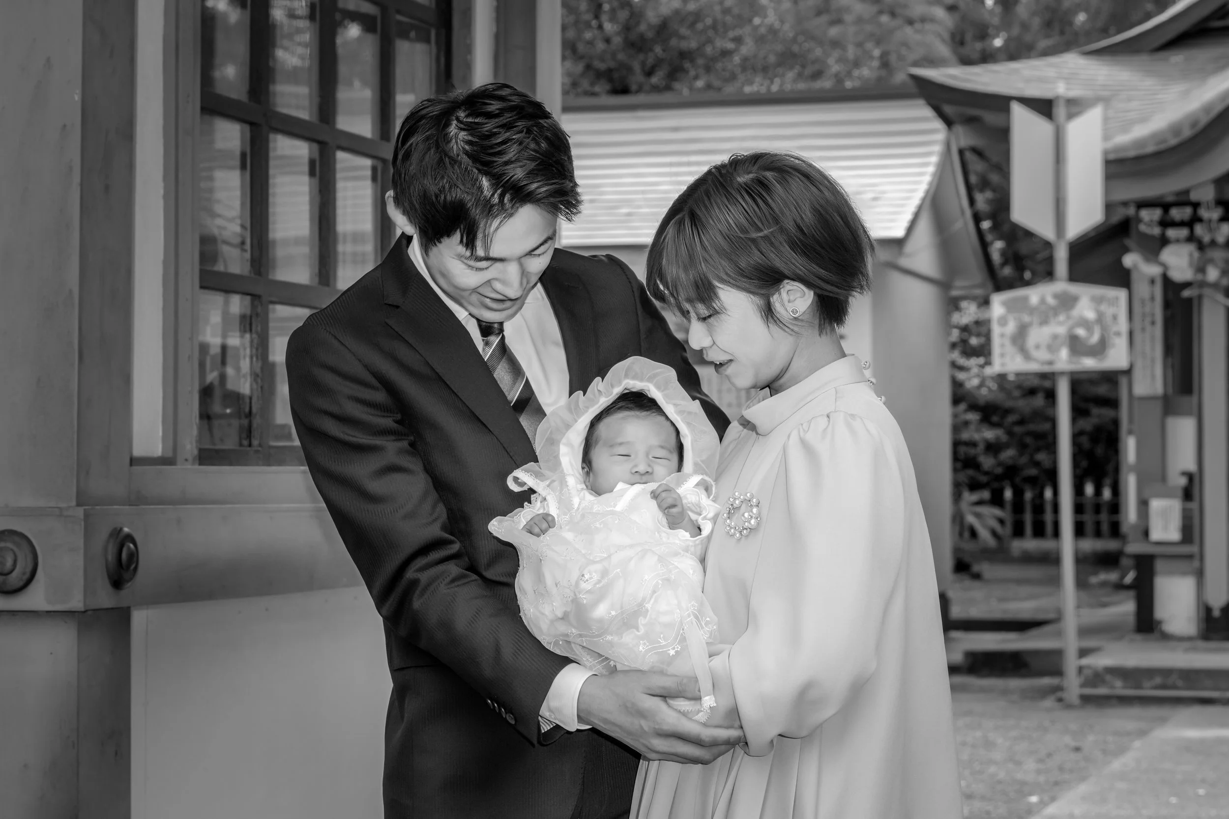 白いドレスを着た赤ちゃんを抱く若いママと、スーツを着た若いパパ。背景に神社のような場所が見える。
