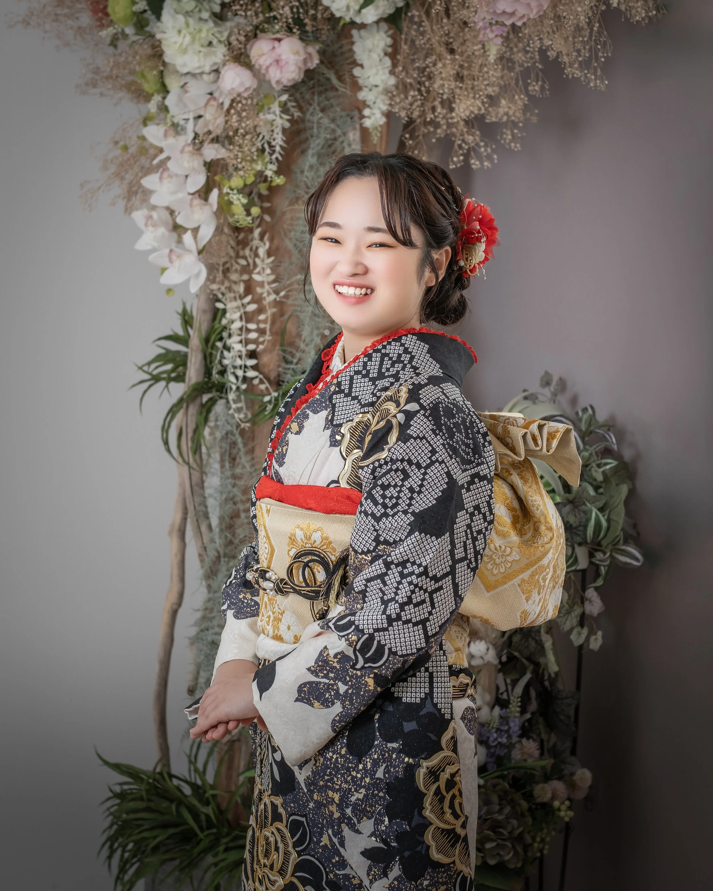 着物を着た笑顔の若い女性がカメラに向かって微笑んでいる。背景には花や植物の装飾がある。