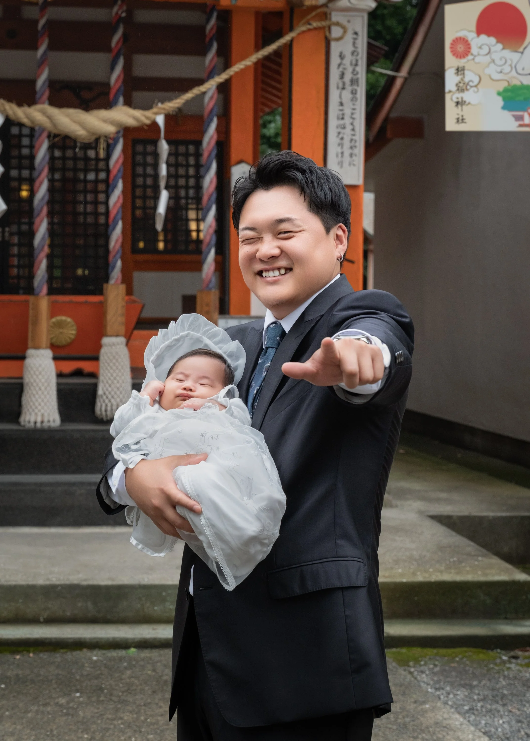 スーツを着た男性が赤ちゃんを抱きながら笑顔で手を差し出している。背景は神社のしめ縄。