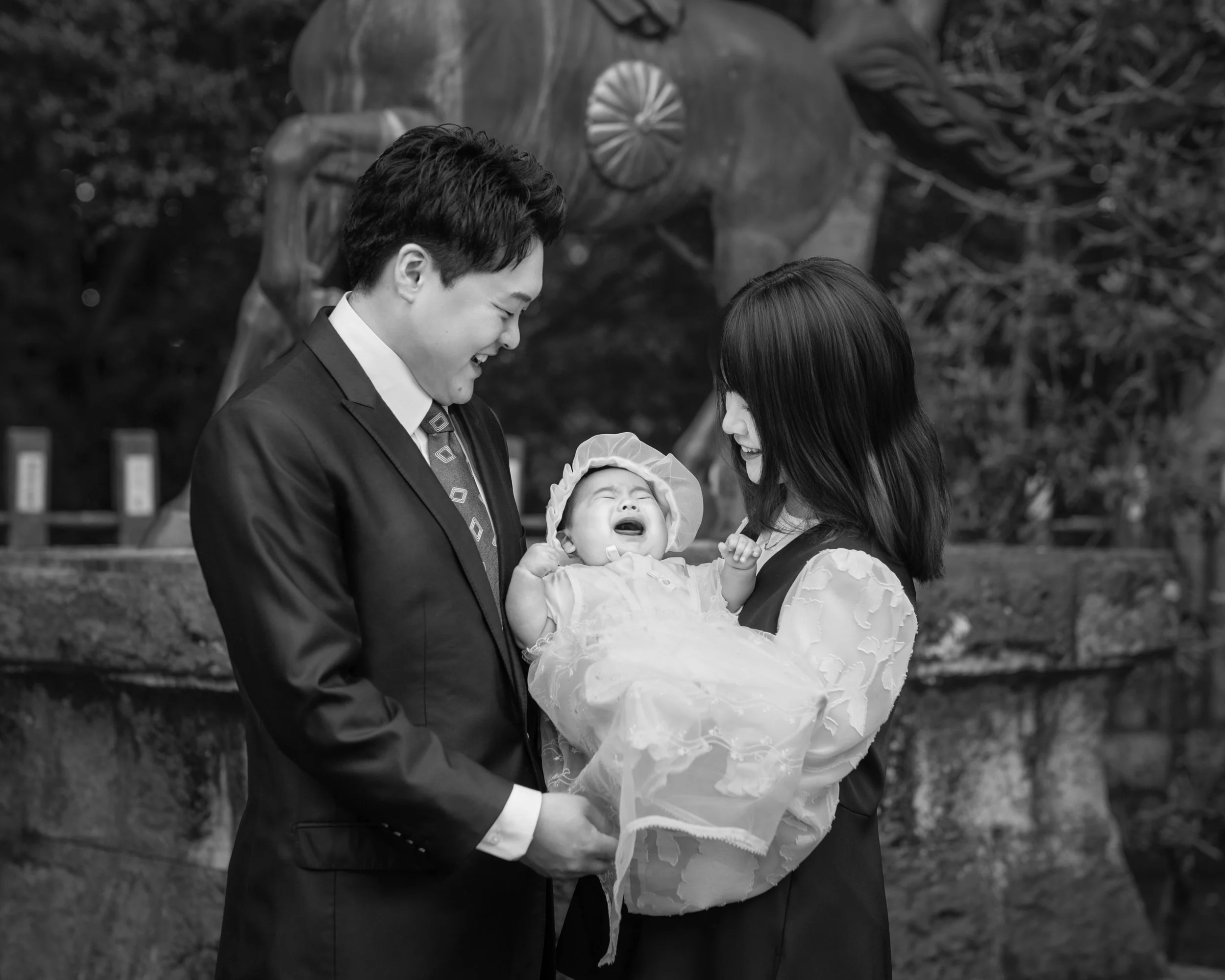 笑顔の両親と泣いている赤ちゃんが木の下にいる。