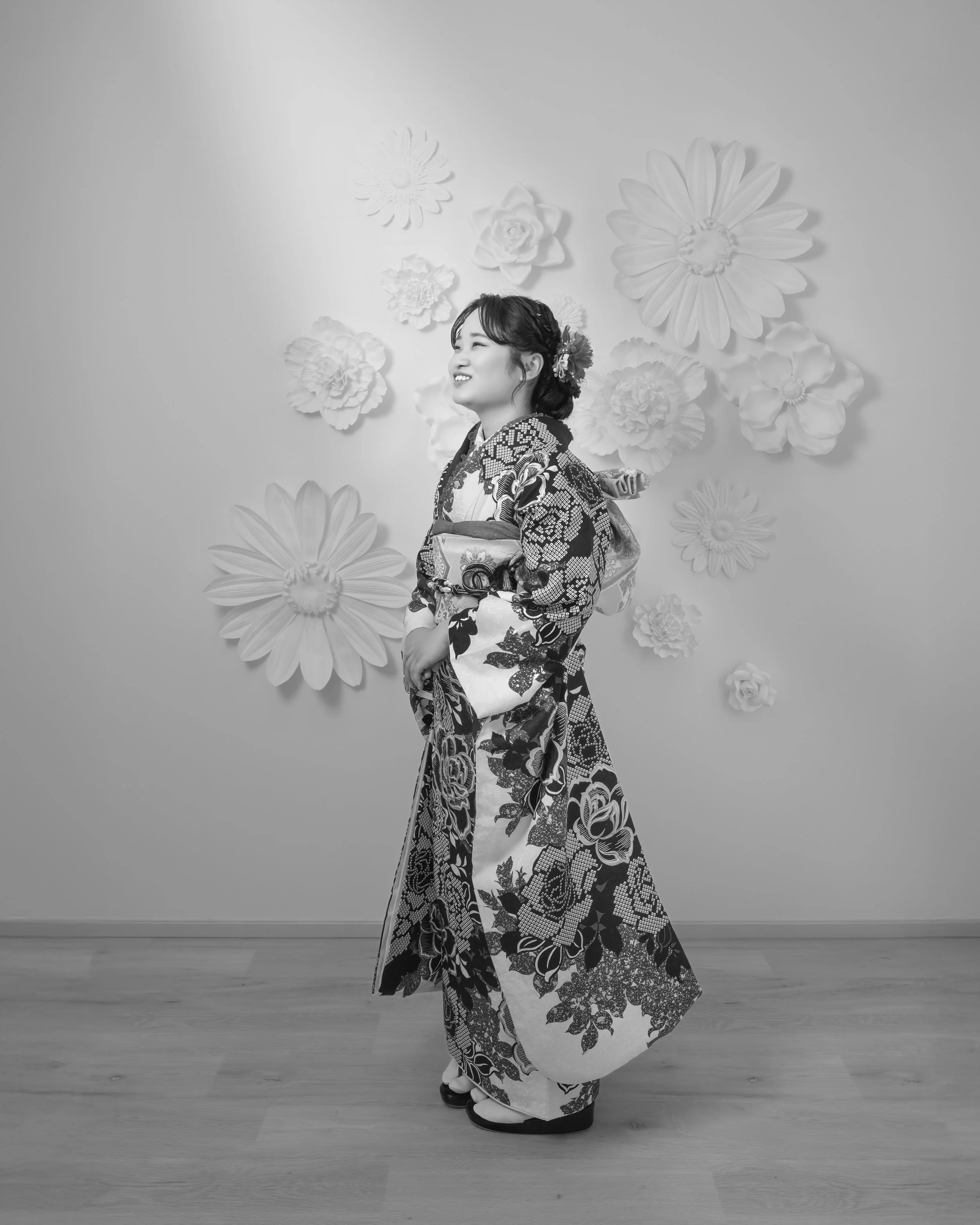 和服を着た女性が笑顔で横を向いている。背景には花の飾りがついた壁がある。