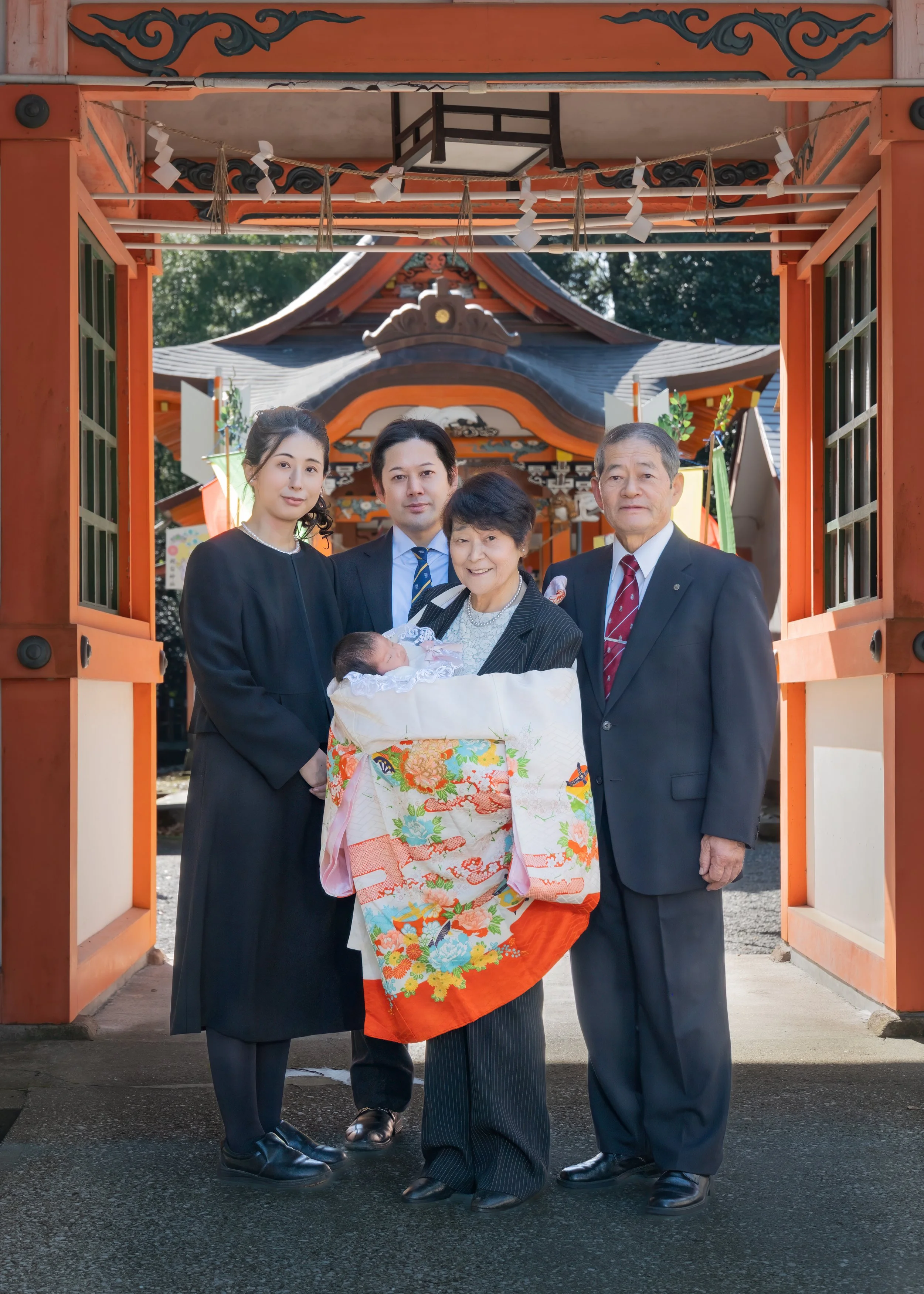 神社の境内で、お宮参りを祝う家族の写真。中央に赤ちゃんを抱く祖母、その周りに若い夫婦と祖父。背景は伝統的な神社の鳥居と建物。