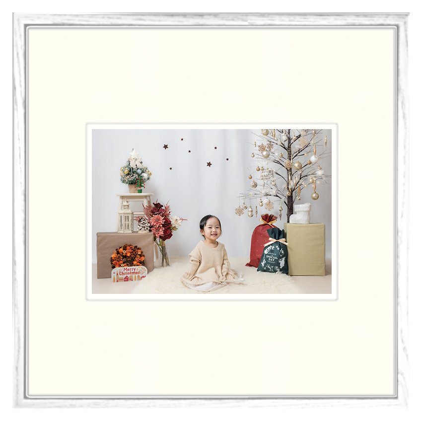 クリスマスの飾り付けの中で笑顔の女の子。ツリーには金色の装飾と白のリボン掛けがされ、周囲にプレゼントと花の飾りが置かれている。