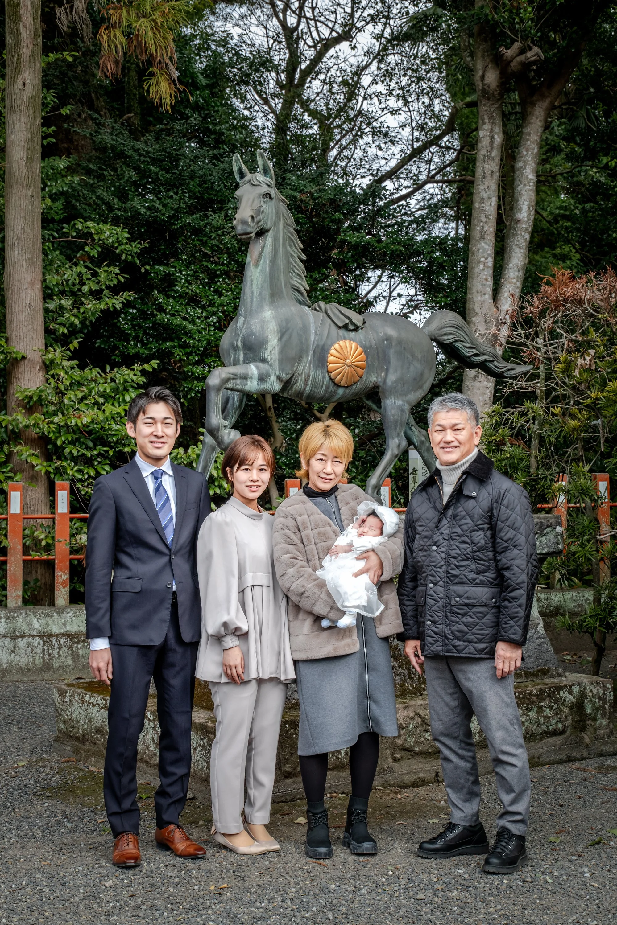 家族が神社の前で写真を撮っています。家族には赤ちゃんを抱いた祖母とその両親、祖父と思われる男性と女性がいます。背景には馬の像があります。