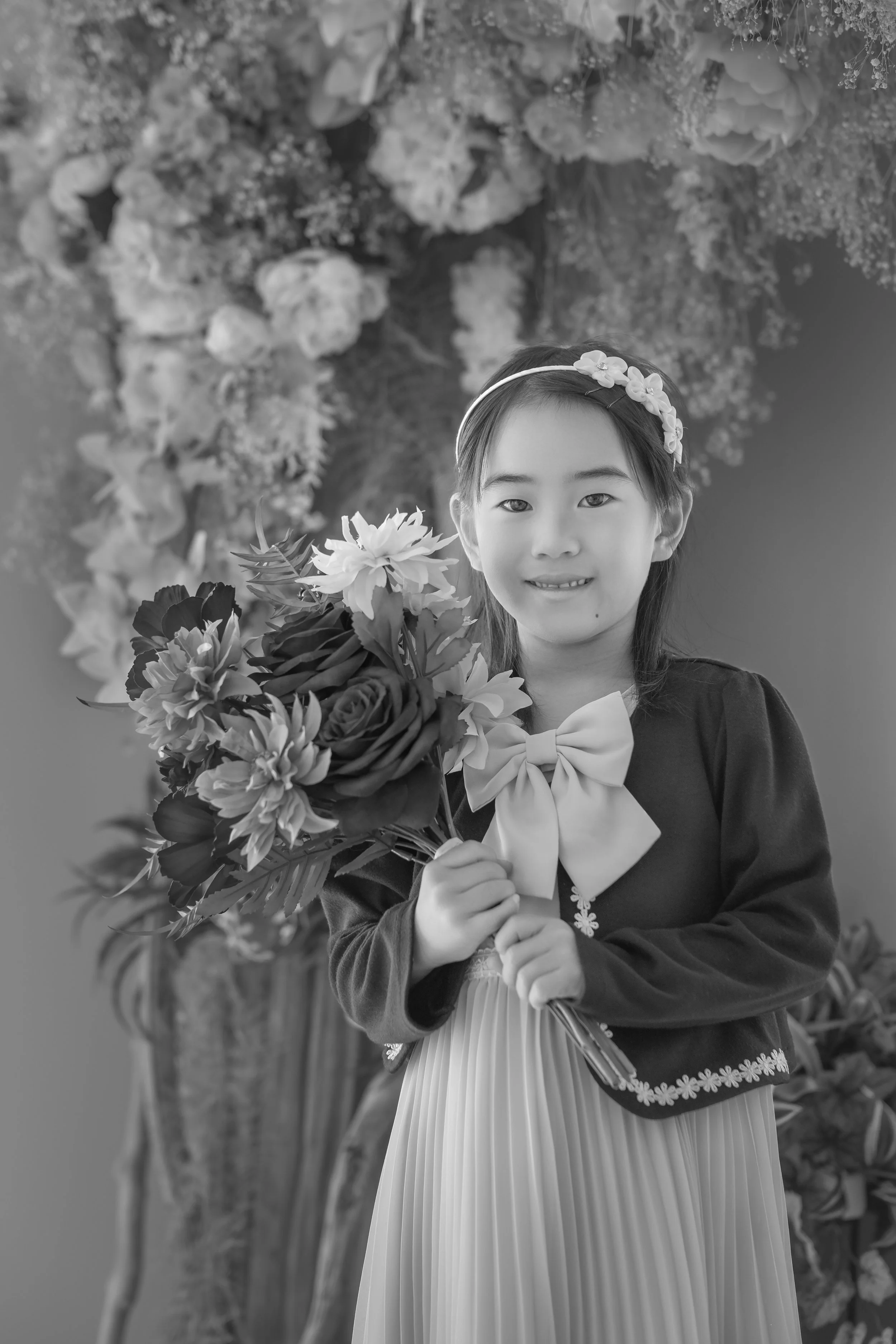 花束を持つ少女が笑顔でポーズをとる写真。背景には花の装飾がある。