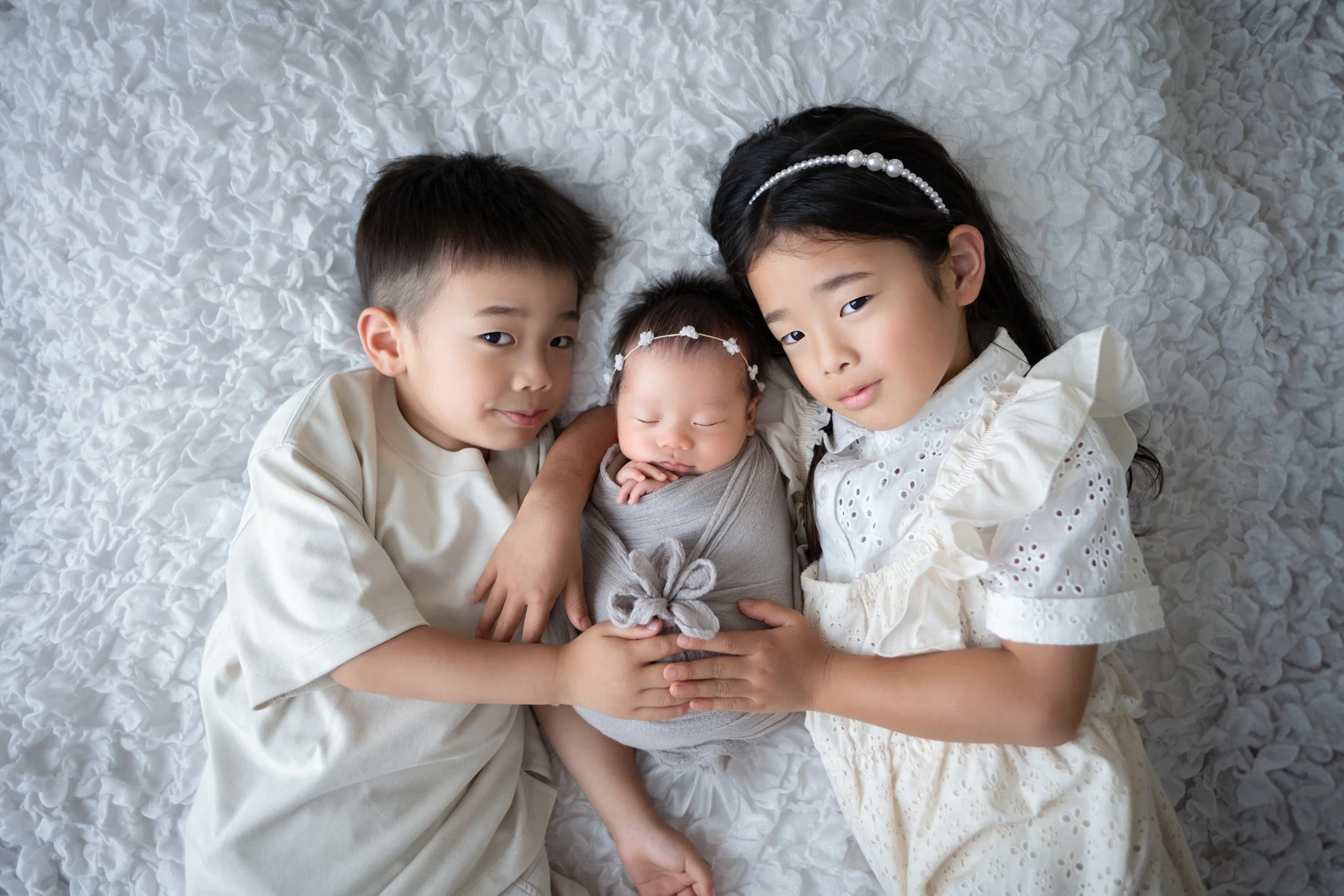 白いシーツの上に寝ている三人の子供。中央は赤ちゃんで、二人の兄妹がそれぞれ左右に位置し、優しく見守っている。