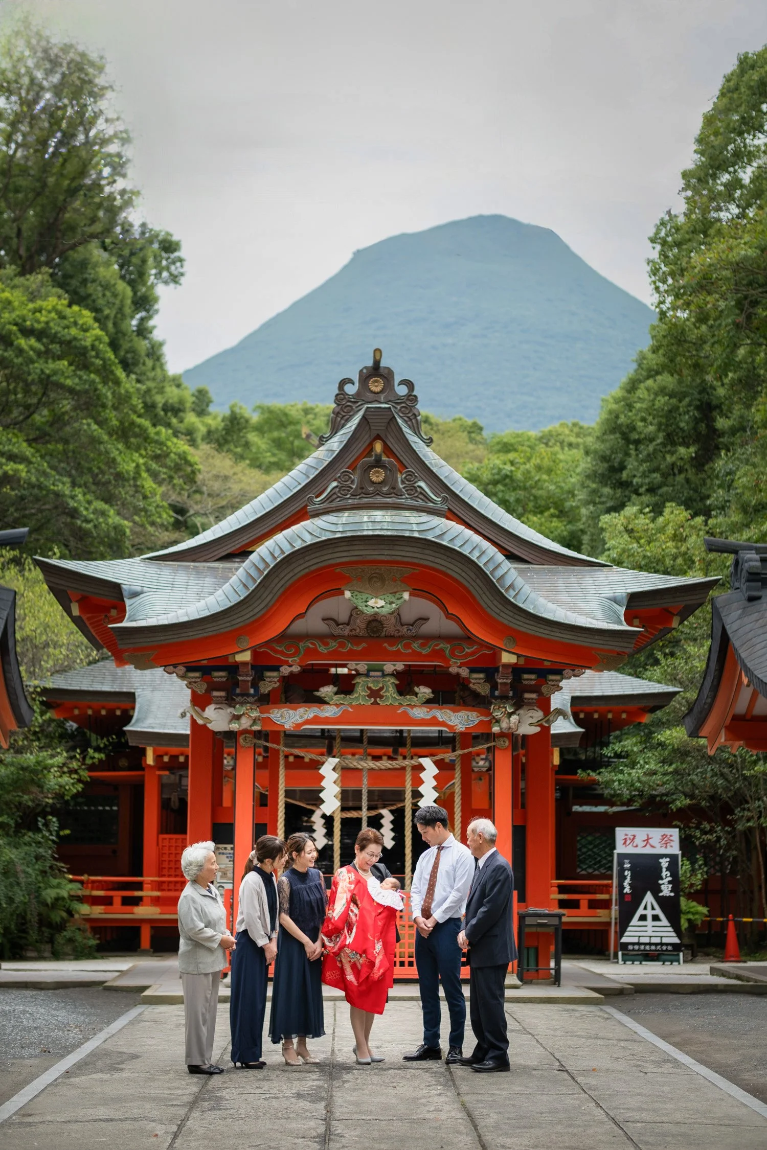 神社の前で新生児の祝福式を行っている家族の様子。背景に緑の木々と山が見える。