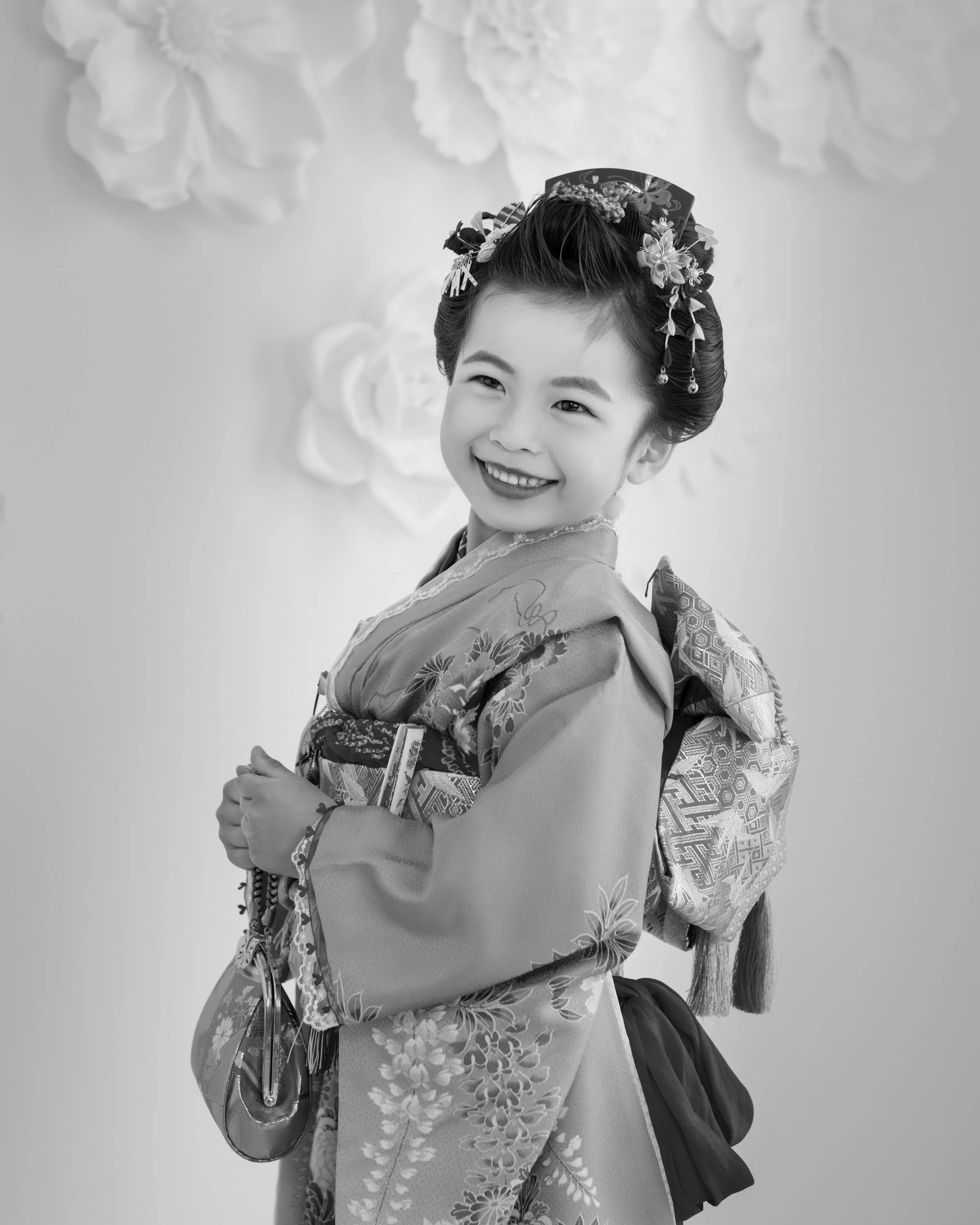 着物を着た少女の黒と白の写真。笑顔で、髪には花飾りをつけている。