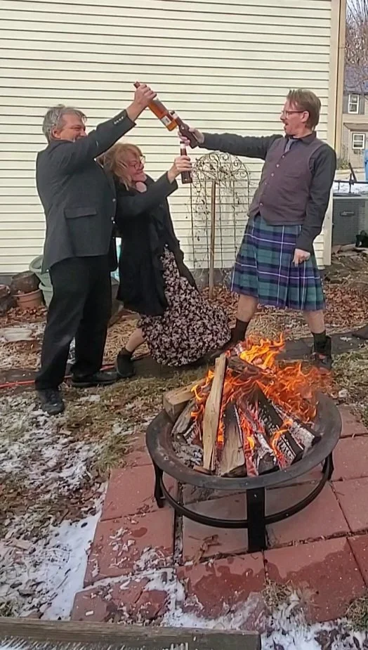 Four people standing around a fire pit outside, cheerfully clinking bottles of beer. The person on the right is wearing a kilt, and the scene appears festive.