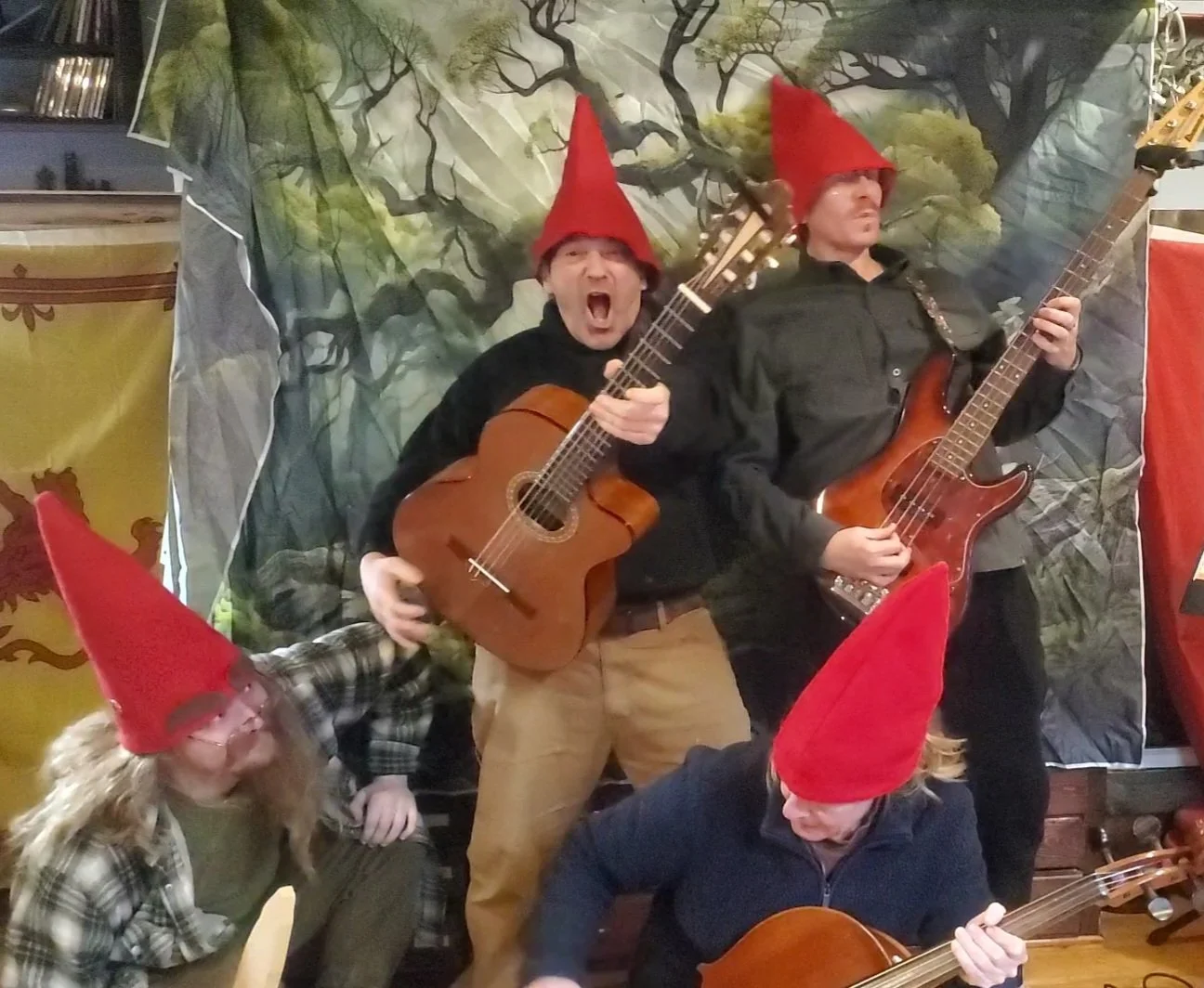 Four people wearing red gnome hats playing musical instruments. Two are playing guitars, one is playing bass, and one is kneeling. The background features a forest scene with trees, and there are decorative banners on either side.
