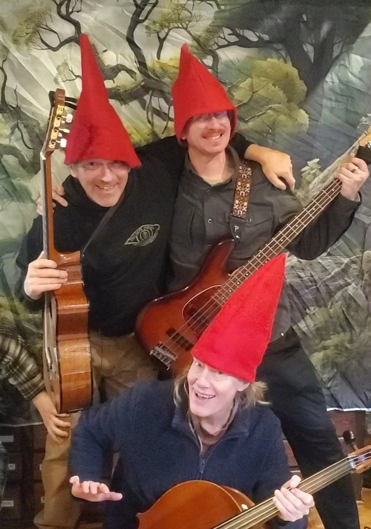 Three people wearing red gnome hats, holding guitars and smiling together, in front of a nature-themed backdrop.