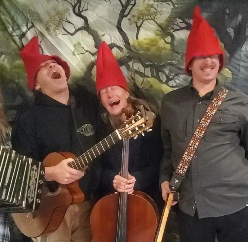 Three people wearing red pointy hats, laughing, with musical instruments, against a forest scene backdrop.