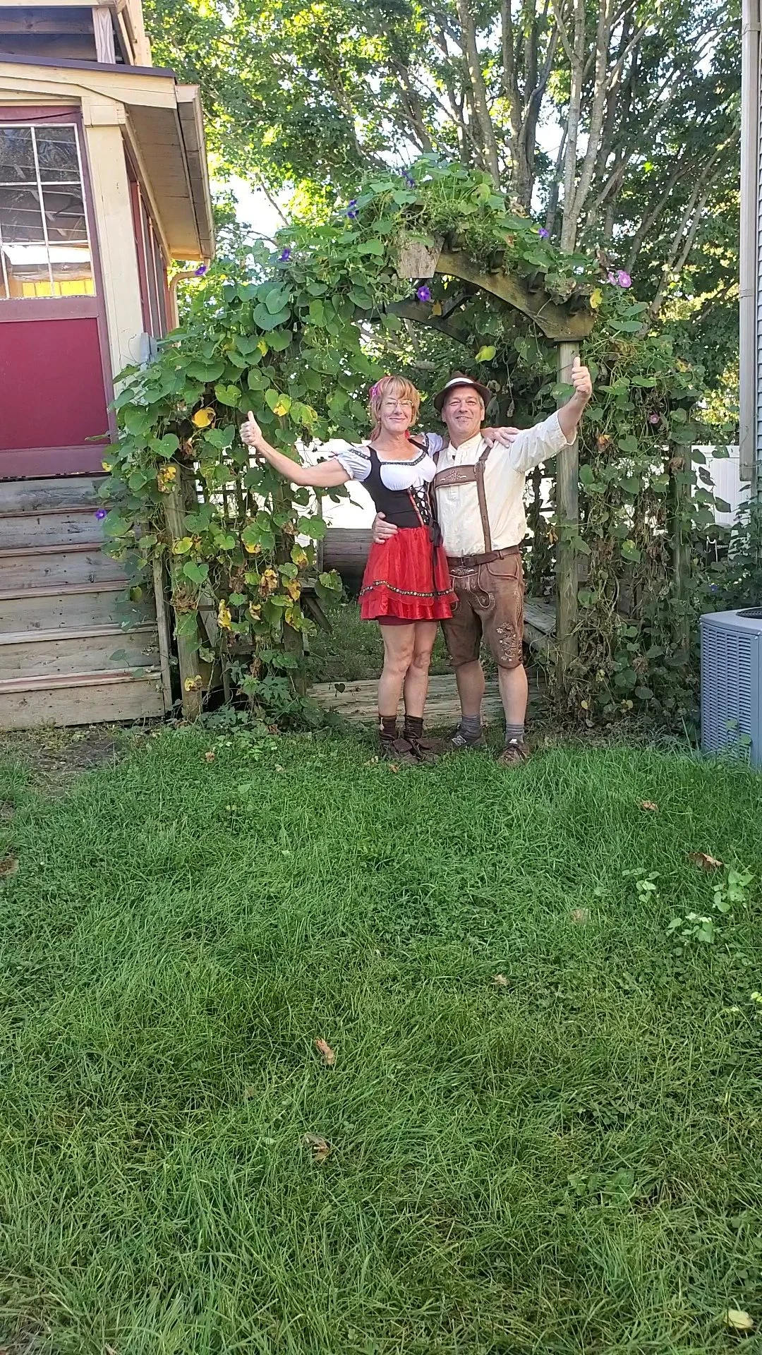 A man and woman dressed in traditional German dirndls and lederhosen, standing in front of a wooden archway with green vines and purple flowers, smiling and giving a thumbs up.
