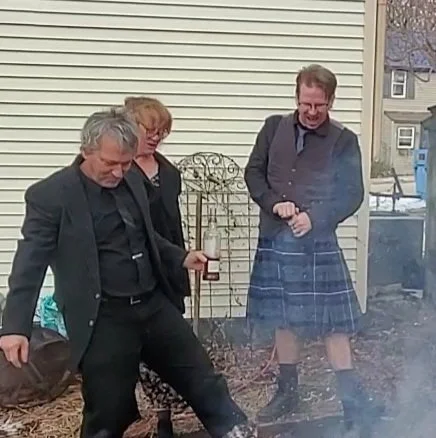 Three people standing outside near a house, two men and one woman, with one of the men holding a beverage can and kicking a fire or barbecue.