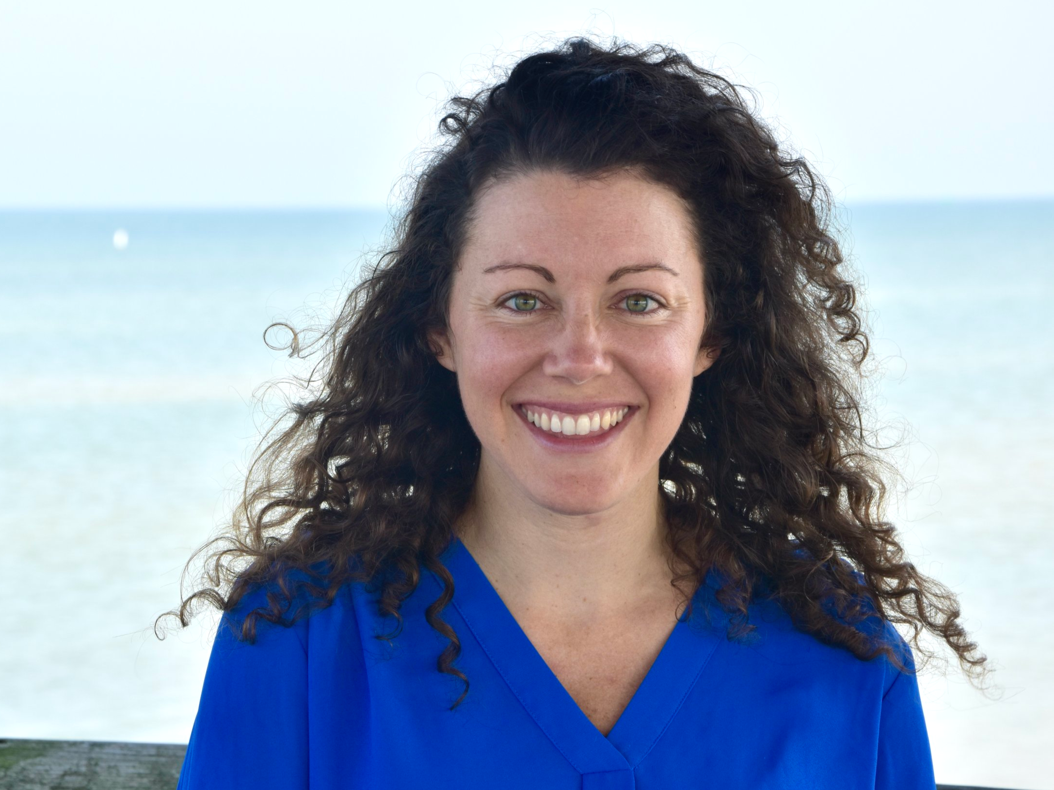 Woman with long curly brown hair and green eyes smiling at the camera, standing outdoors near the beach in a blue top.