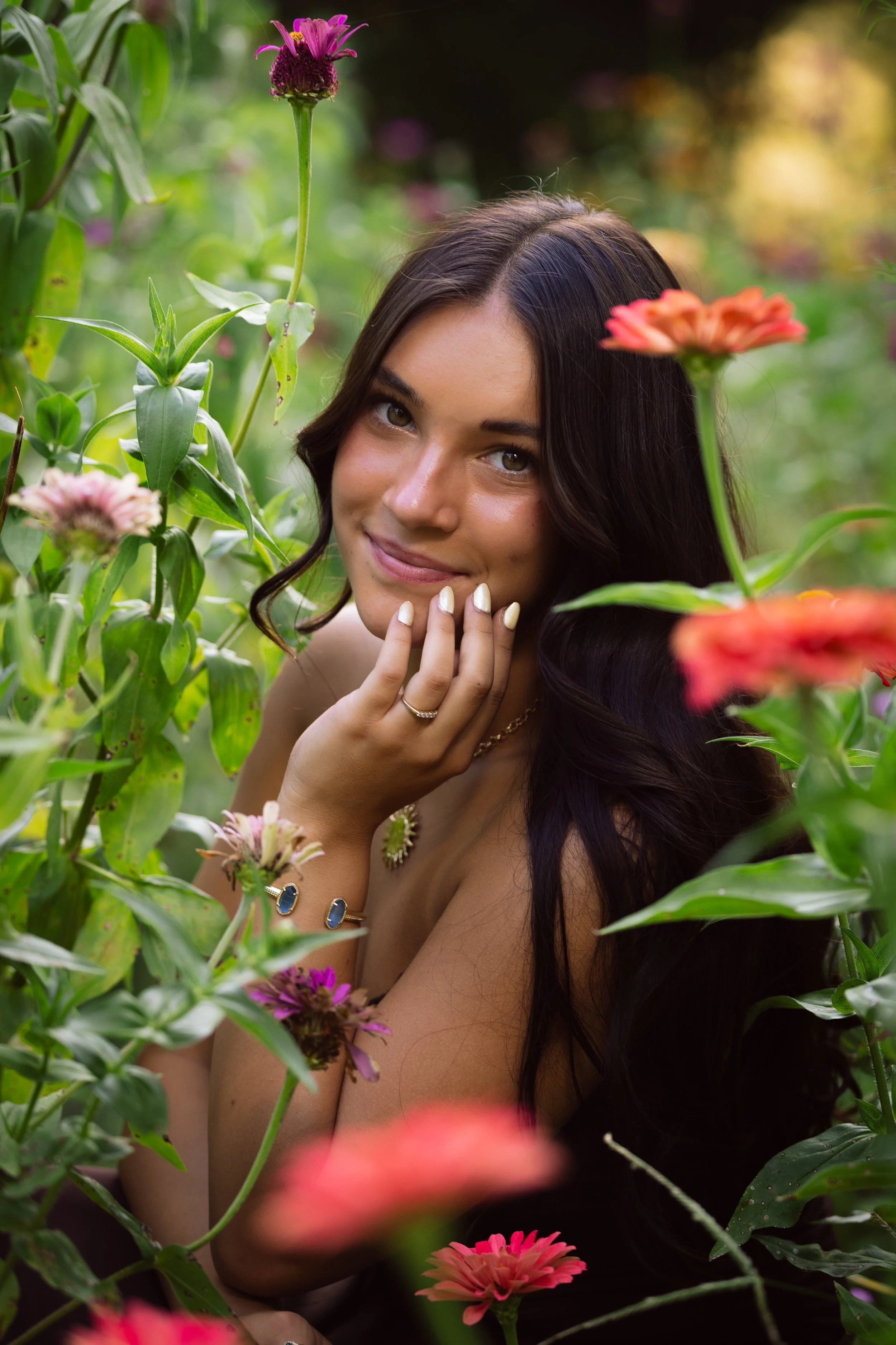 A woman with long dark hair smiling while surrounded by colorful flowers and green foliage in a natural outdoor setting. Senior Class of 2027 portrait photographer best photographer in northwest arkansas