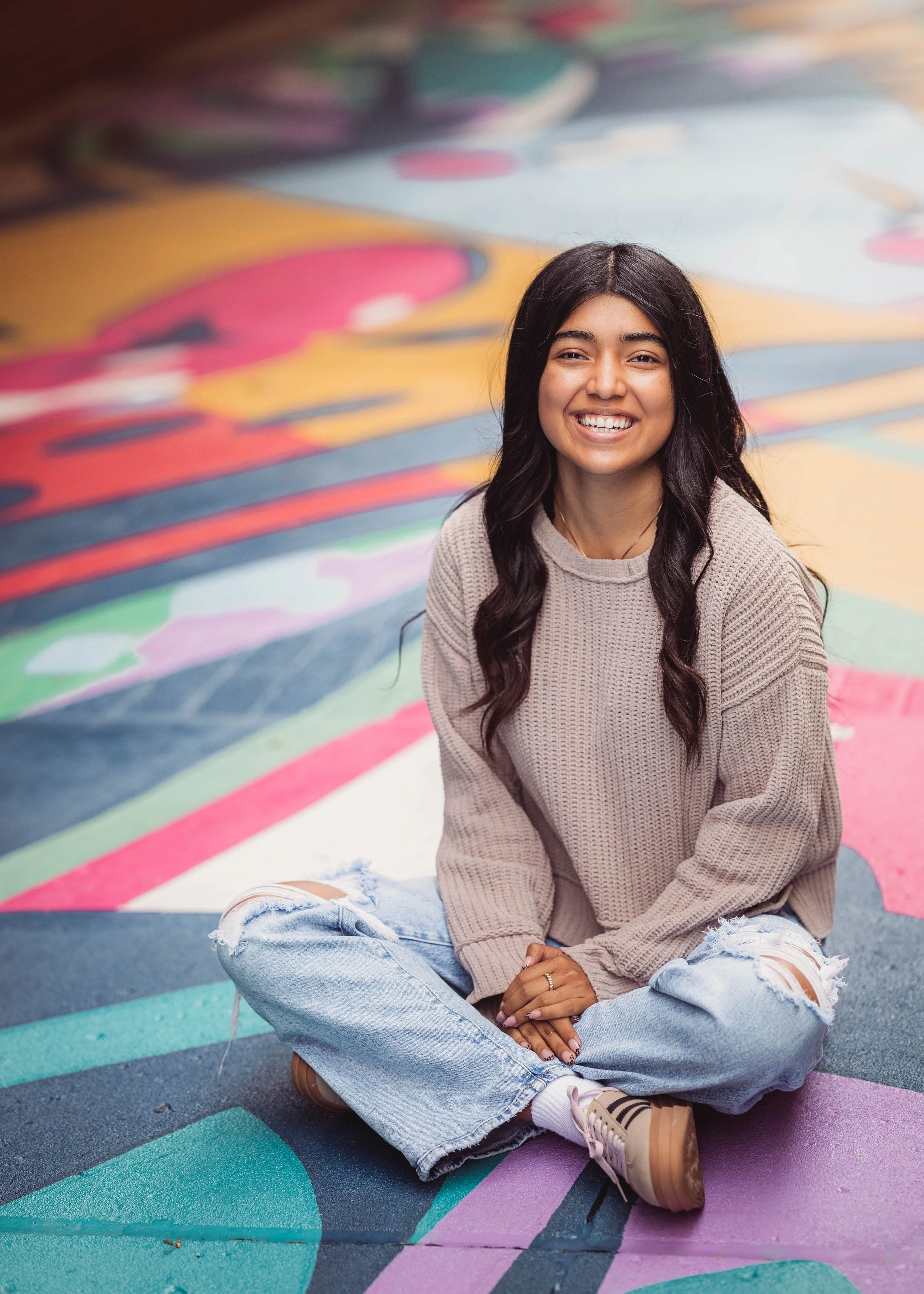 A young woman with long dark wavy hair, smiling and sitting cross-legged on a colorful, abstract painted ground.