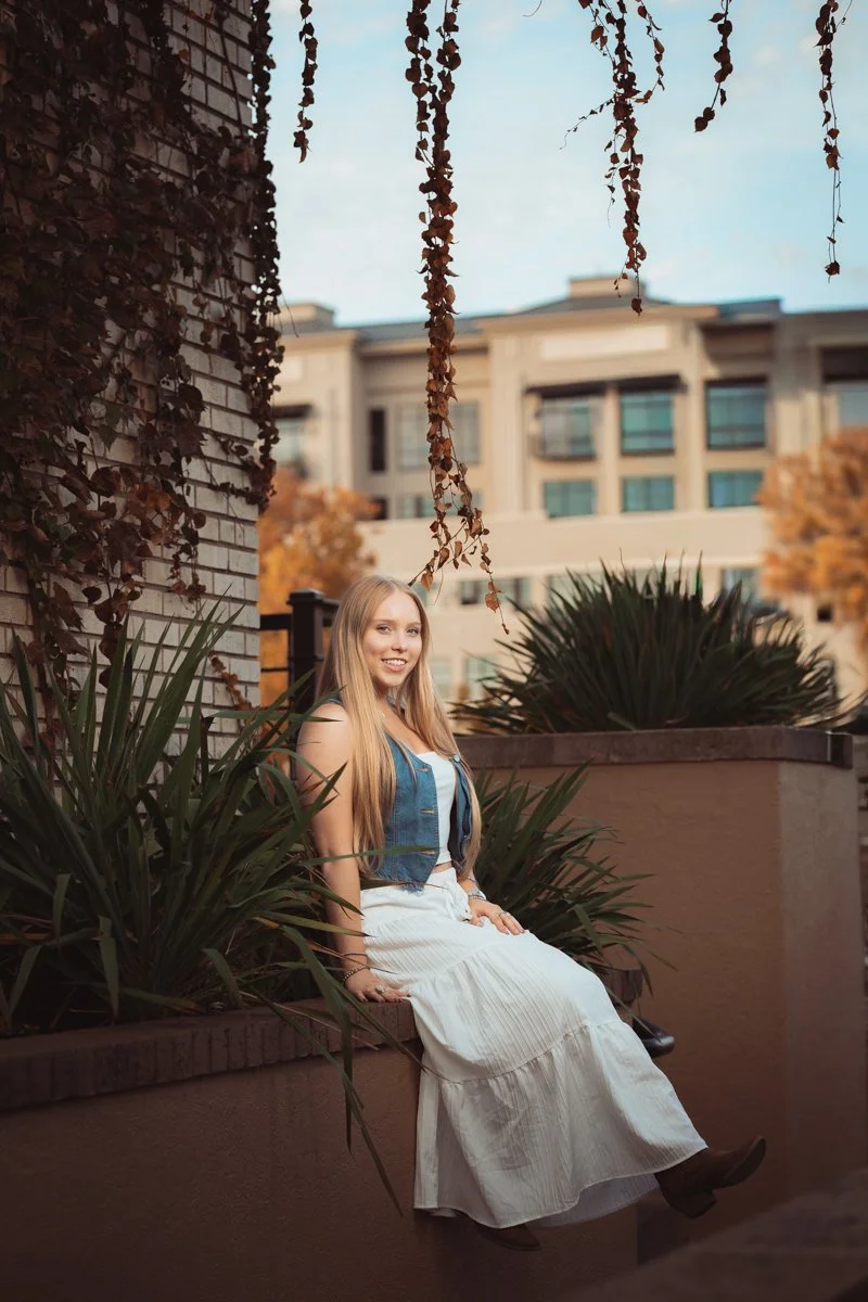 A young woman with long blonde hair sitting on a brick wall, surrounded by plants, with a large apartment building in the background during daytime.