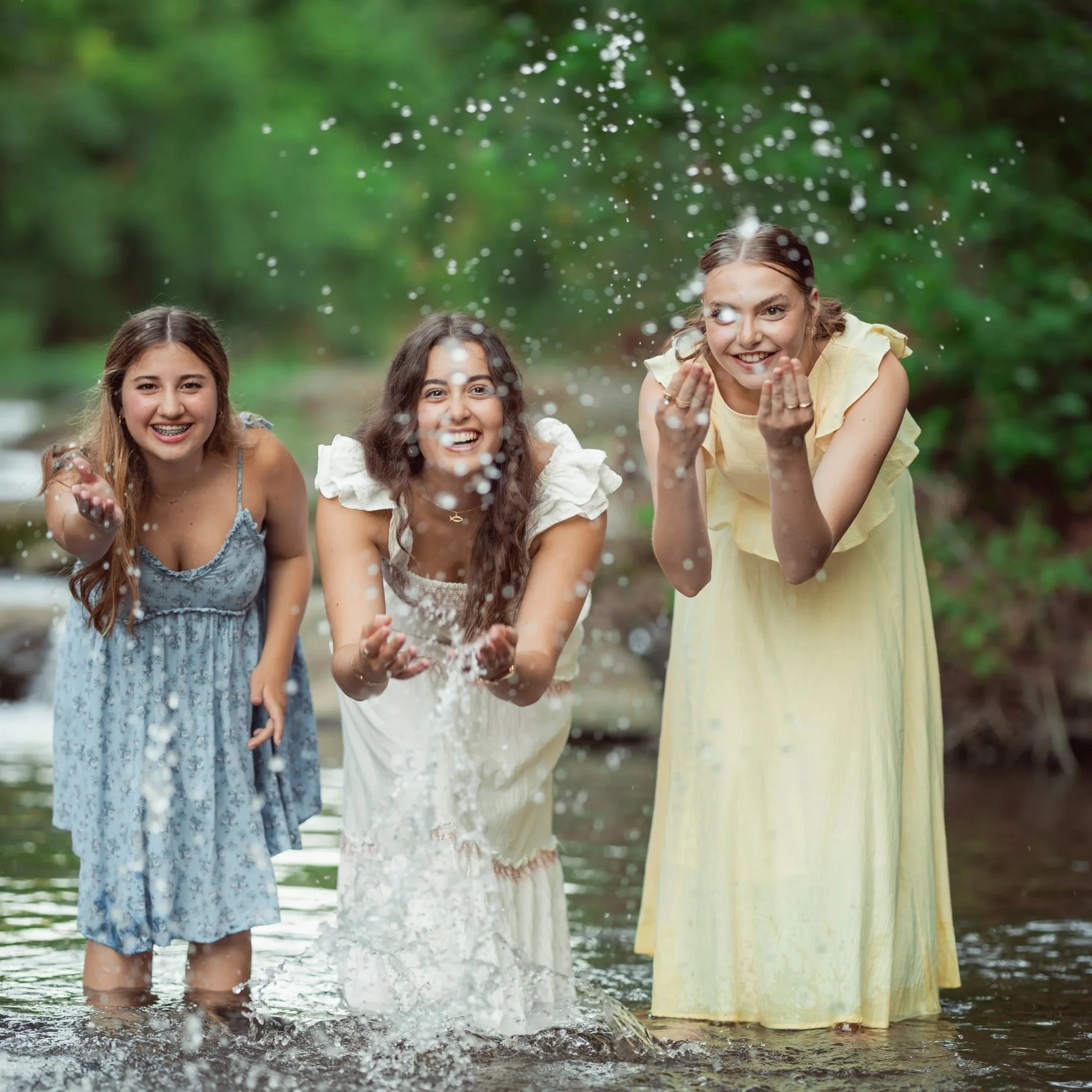 Three young women in summer dresses playing and splashing water in a shallow stream in a lush green outdoor setting, smiling and enjoying themselves.