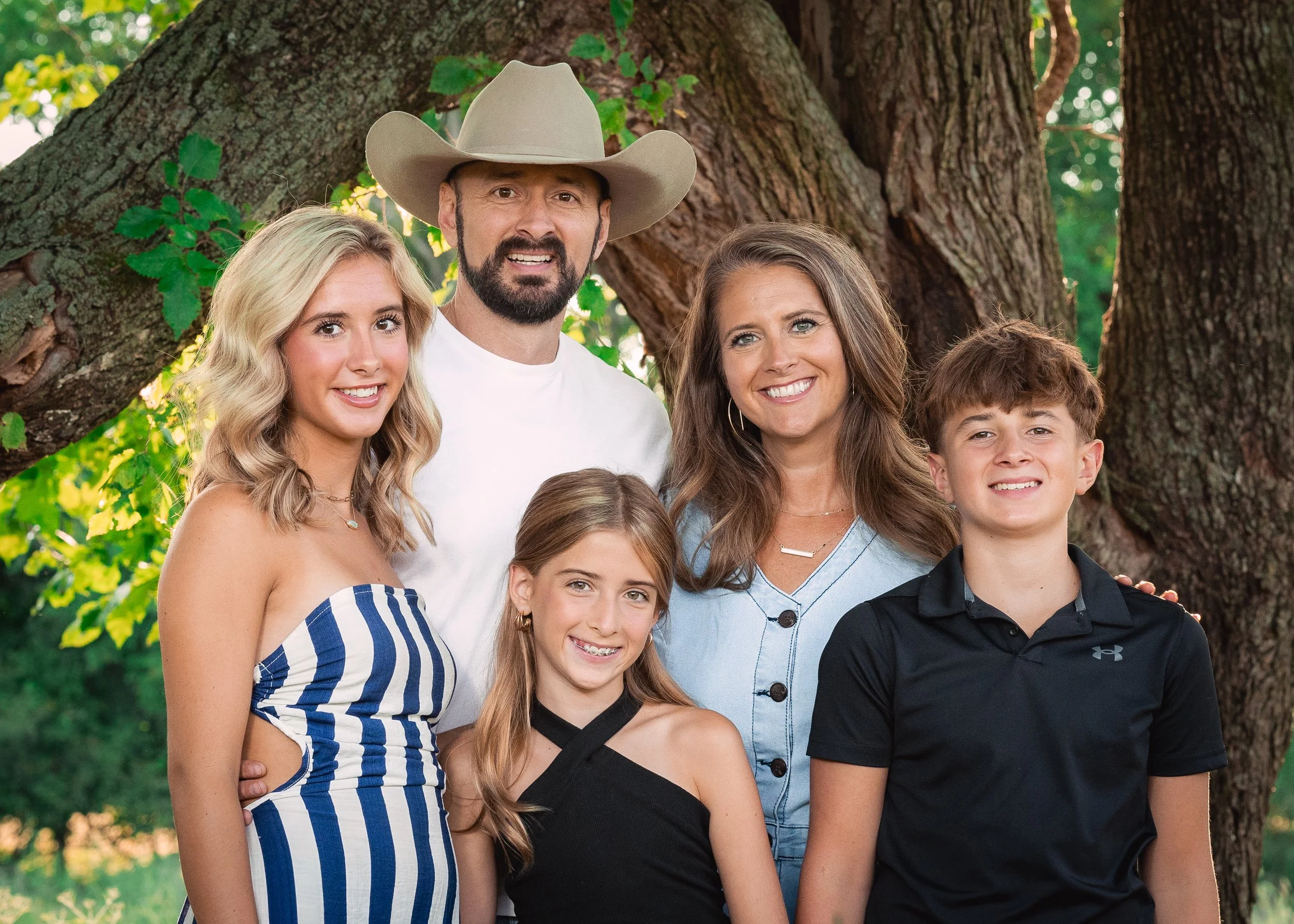 A family of six posing outdoors near a large tree, smiling and dressed casually in summer clothing.