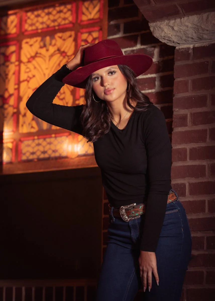 A woman with wavy dark hair, wearing hoop earrings, a black long-sleeve shirt, dark jeans, a decorative belt, and a wide-brimmed maroon hat, standing against a brick wall with a colorful stained glass window in the background.