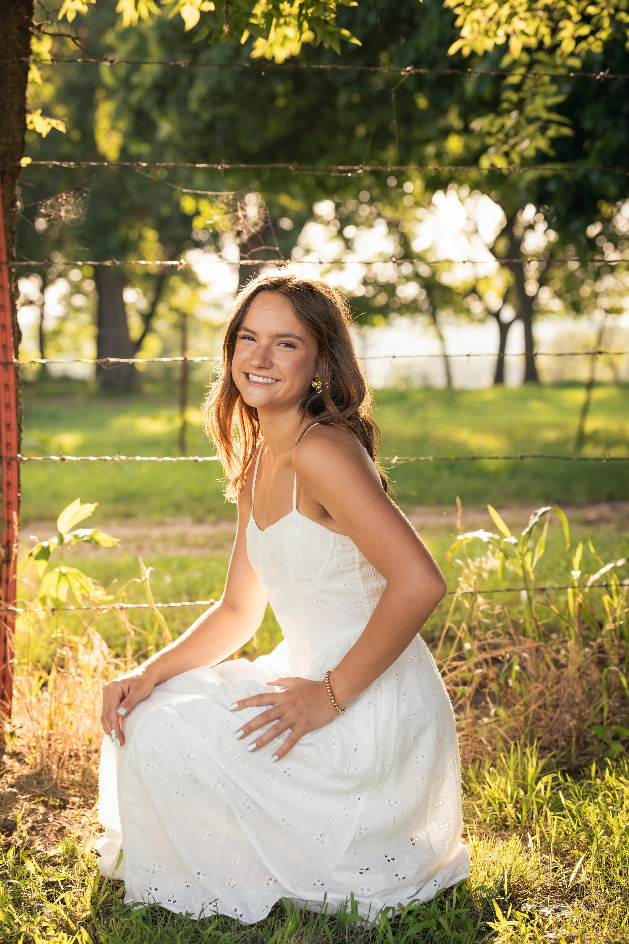 Smiling woman in a white dress sitting outdoors near a barbed wire fence, with green trees and sunlight in the background.