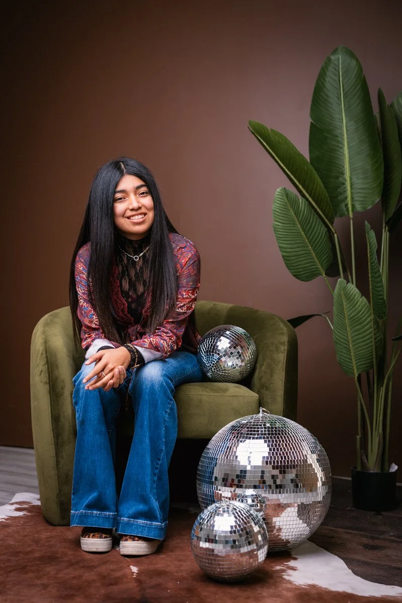 A young woman with long black hair sitting on a green velvet armchair, smiling at the camera, with three disco balls of varying sizes on the floor and on the chair, and a tall green plant with large leaves in the background against a brown wall.