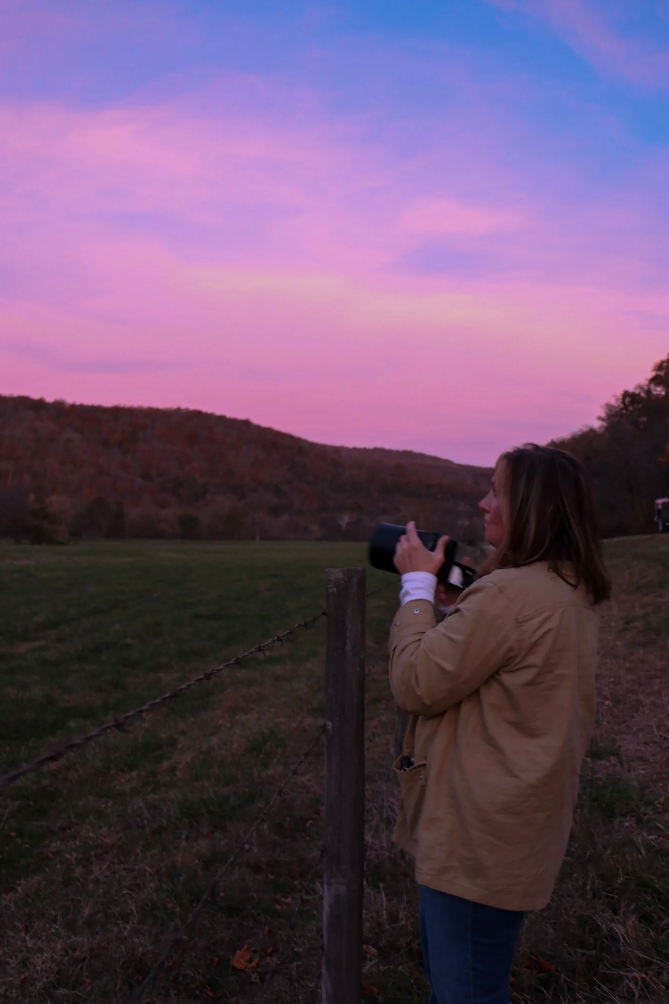 A woman with shoulder-length hair, wearing a tan jacket and blue jeans, stands outdoors behind a wooden fence, holding a camera with a large zoom lens, capturing a scenic sunset with pink and purple clouds over rolling hills.